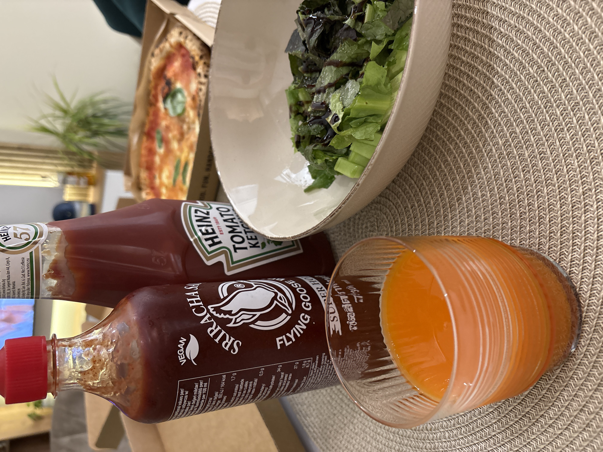 The image displays an indoor setting, likely a dining area in Sofia, Bulgaria, featuring a casual meal laid out on a light-colored woven placemat.
In the background, a portion of a pizza slice is visible within its cardboard box, topped with a red sauce and fresh basil leaves. Beside the pizza, a light-colored ceramic bowl contains a green salad, consisting mainly of lettuce with a dark dressing drizzled over it.
Two condiment bottles are prominently placed on the placemat. One is a classic red Heinz Tomato Ketchup bottle, displaying its iconic "HEINZ", "TOMATO KETCHUP", and "57" labels, along with "ESTP 1869". The label also includes distribution information for "Bucuresti, Romania" and "Lefkosa" (Nicosia), and states "Polonya" (Poland) as the country of manufacture, alongside nutritional values like "693 kJ/164 kcal" per 100 ml. The other bottle is a brown Flying Goose Sriracha Hot Chili Sauce, marked "VEGAN", with "SRIRACHA" and "FLYING GOOSE" clearly visible. This bottle also presents nutritional information in multiple languages, including Finnish and Turkish, indicating broad distribution.
Next to the condiment bottles, a glass with a distinctive ribbed texture holds an orange-colored beverage, likely juice. Visible text on the glass includes "SUFFER HO" and "730". In the blurred background, a small green houseplant and a reflection from what appears to be a television or screen can be seen. The overall scene suggests a relaxed meal at home.