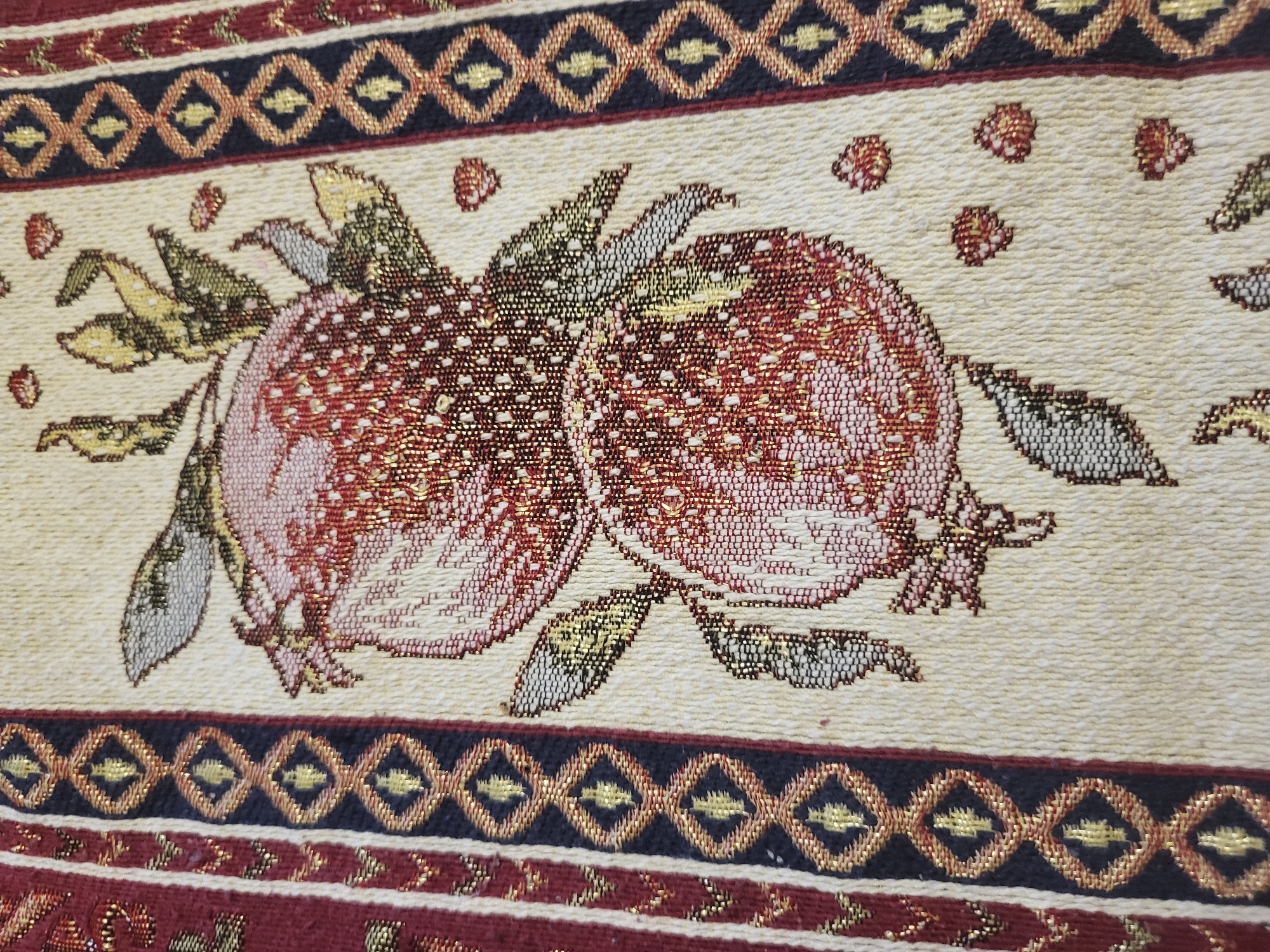 The image shows a close-up of a woven tapestry or tablecloth with a fruit motif. Two large, round fruits, likely pomegranates, dominate the central design. They are primarily red and pink, with detailed woven texture. Leaves in shades of green, gray, and yellow are visible around the fruits. The background is a light beige or cream color, with small red flower-like accents scattered throughout. A decorative border runs along the top and bottom edges of the image. It features alternating dark blue and red bands with a diamond and geometric pattern in gold. The style of the tapestry suggests a possible origin in Armenia, given the prominence of pomegranate motifs in Armenian art and culture, and the style is consistent with traditional Armenian weaving. However, without additional information, this association with Yerevan or any specific city location is speculative.