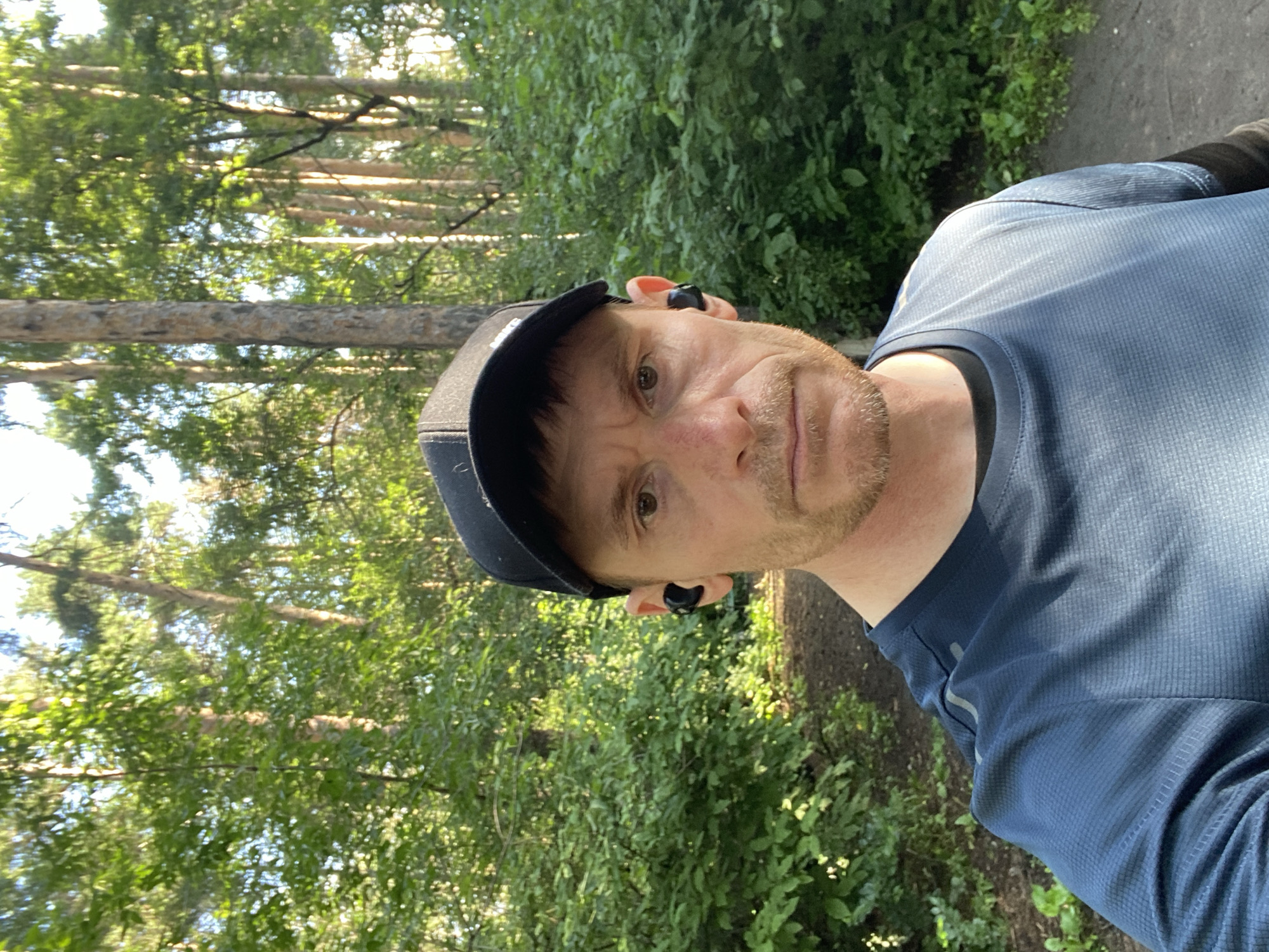 A medium shot selfie shows a Caucasian man in his late 30s or early 40s wearing a dark baseball cap and a blue long-sleeved shirt. He has short brown hair, a light beard and mustache, and is wearing black wireless earbuds. He appears to be outdoors in a wooded area with a dirt path visible behind him. The trees are tall and covered in green leaves, suggesting it is summer. The lighting indicates it is daytime, likely late afternoon or early evening, given the angle of the sun casting shadows. The man is looking directly at the camera with a neutral expression. The scene is consistent with a park or forest trail in Yekaterinburg, Russia, during the warmer months.