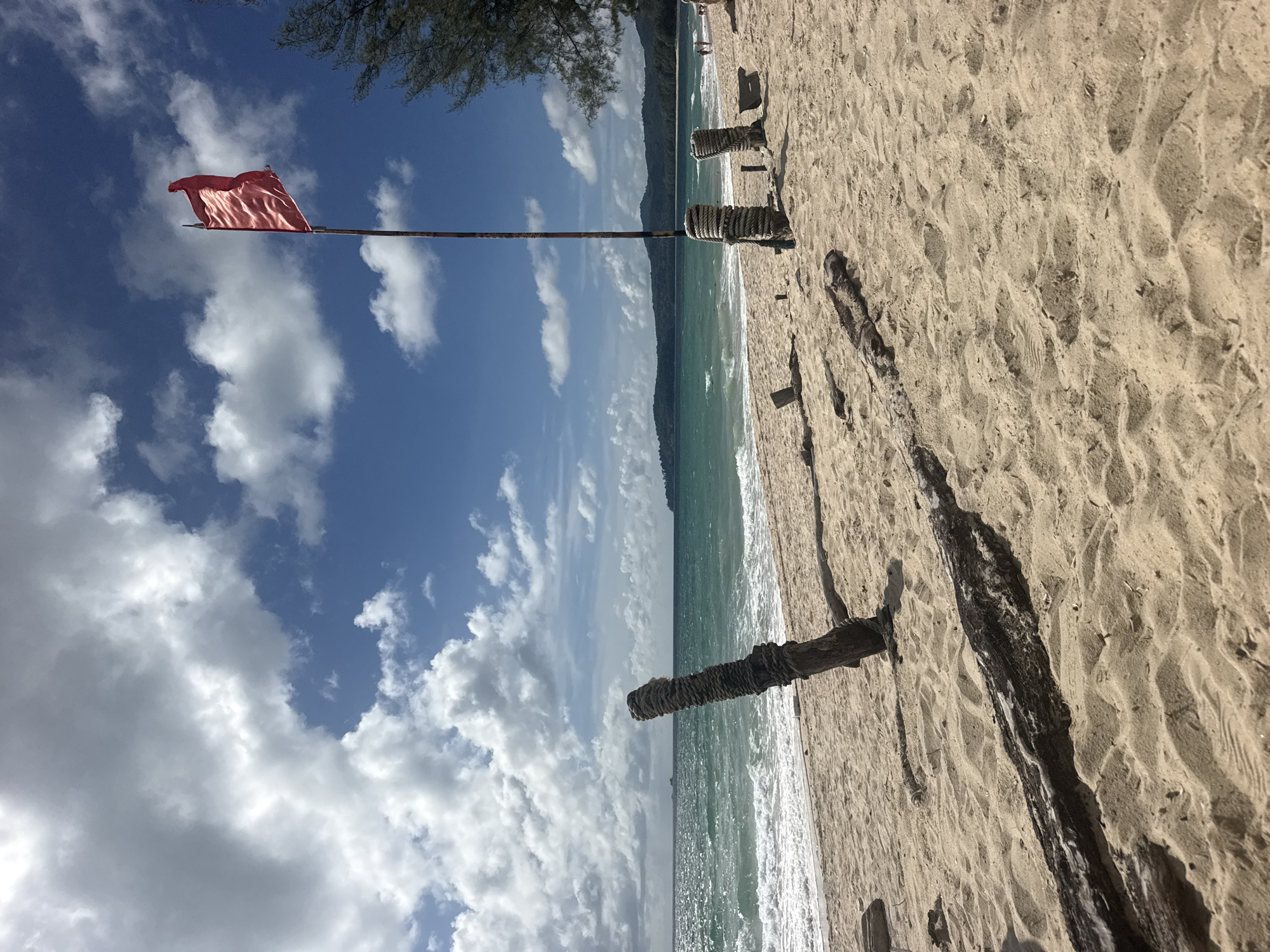 The image depicts a tropical beach scene in Choeng Thale, Thailand. The foreground is dominated by the sandy beach, with several pieces of driftwood scattered across it. In the midground, the turquoise ocean meets the shore with gentle waves, and a red flag flies atop a tall pole. The background features a lush, green coastline with hills and mountains, under a bright blue sky with scattered white clouds. A few people are visible walking along the shoreline in the distance. The overall atmosphere is serene and inviting, suggesting a perfect day for relaxation and outdoor activities.