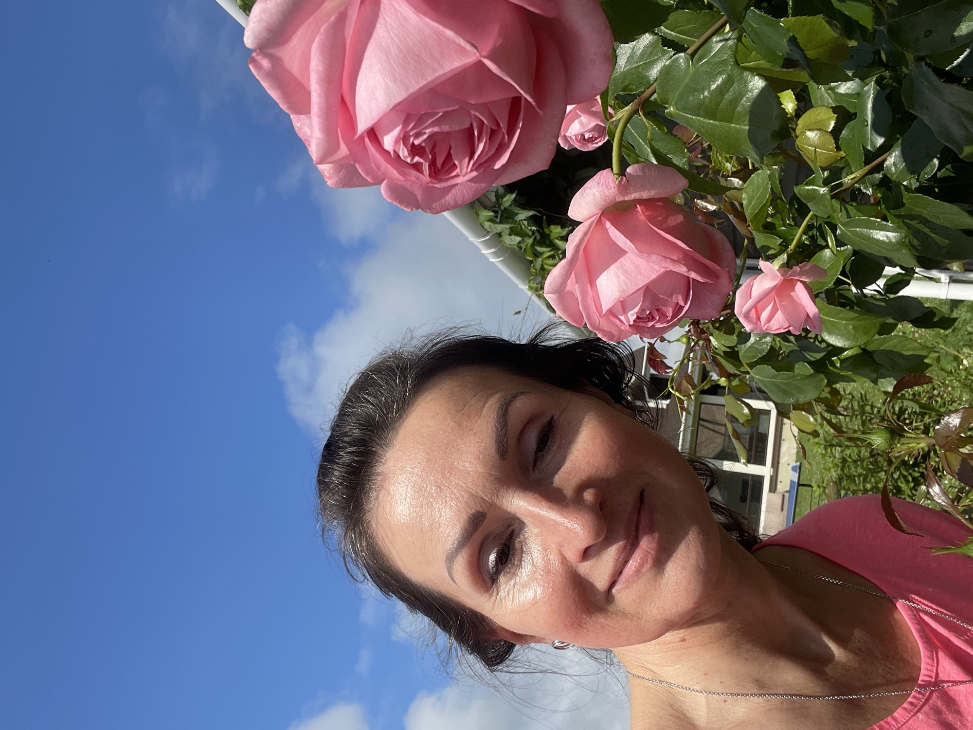 A woman is taking a selfie in front of a rose bush with pink roses. She is wearing a pink t-shirt and a silver necklace. The sky is blue with some white clouds. In the background, there is a house with windows and a garden. The image appears to be taken in the city of Solov'evka, Russia.