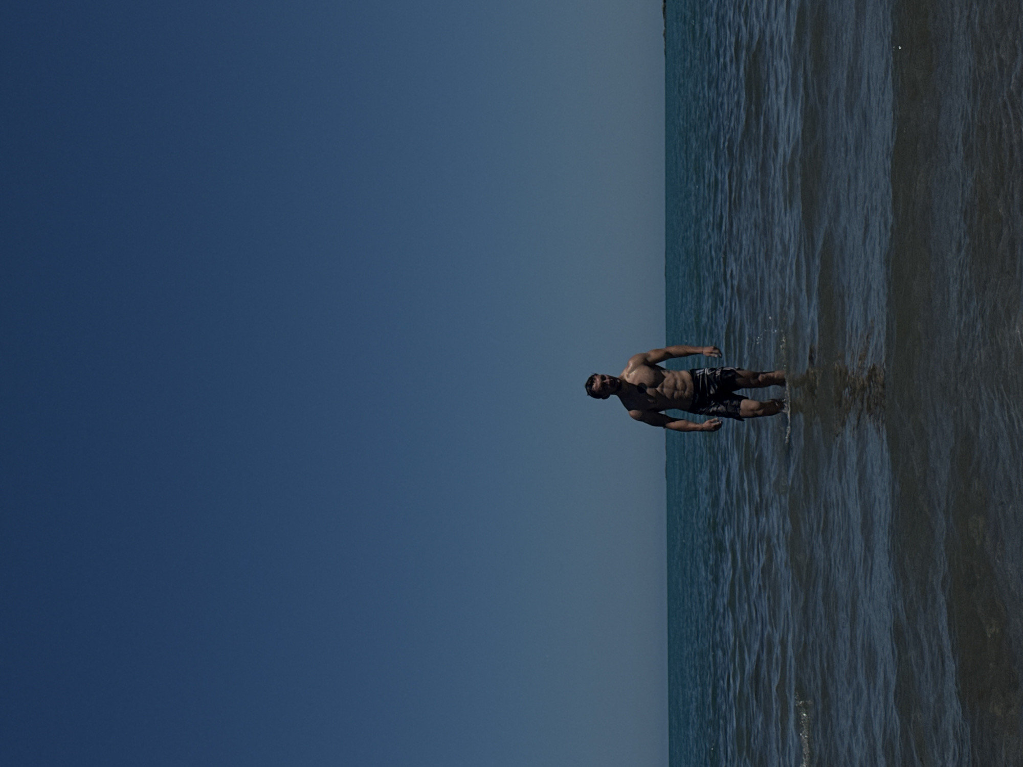 A man with a muscular physique stands in knee-deep water under a clear blue sky. He is wearing dark-colored swim trunks with a patterned design. The water is calm with gentle ripples, and the horizon where the sea meets the sky is visible in the background. The scene appears to be a beach or coastal area. The overall impression is one of a serene, sunlit day at the seaside.