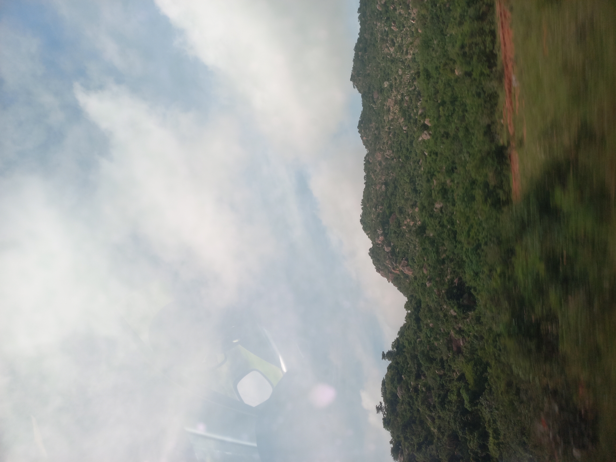 The image depicts a landscape dominated by a verdant, rocky hillside under a sky filled with white, scattered clouds. The foreground is blurred due to motion, showing streaks of green foliage and patches of reddish-brown earth. The hillside itself is densely covered with trees and shrubs, with visible grey rocks and boulders interspersed throughout. The sky is bright and partially obscured by clouds, suggesting it is daytime. Reflections are visible in the upper left quadrant, indicating the photograph was taken from inside a vehicle or structure with windows. There are no people or animals visible in the scene. The location is specified as Soka, a district in Ibadan, Oyo State, Nigeria.