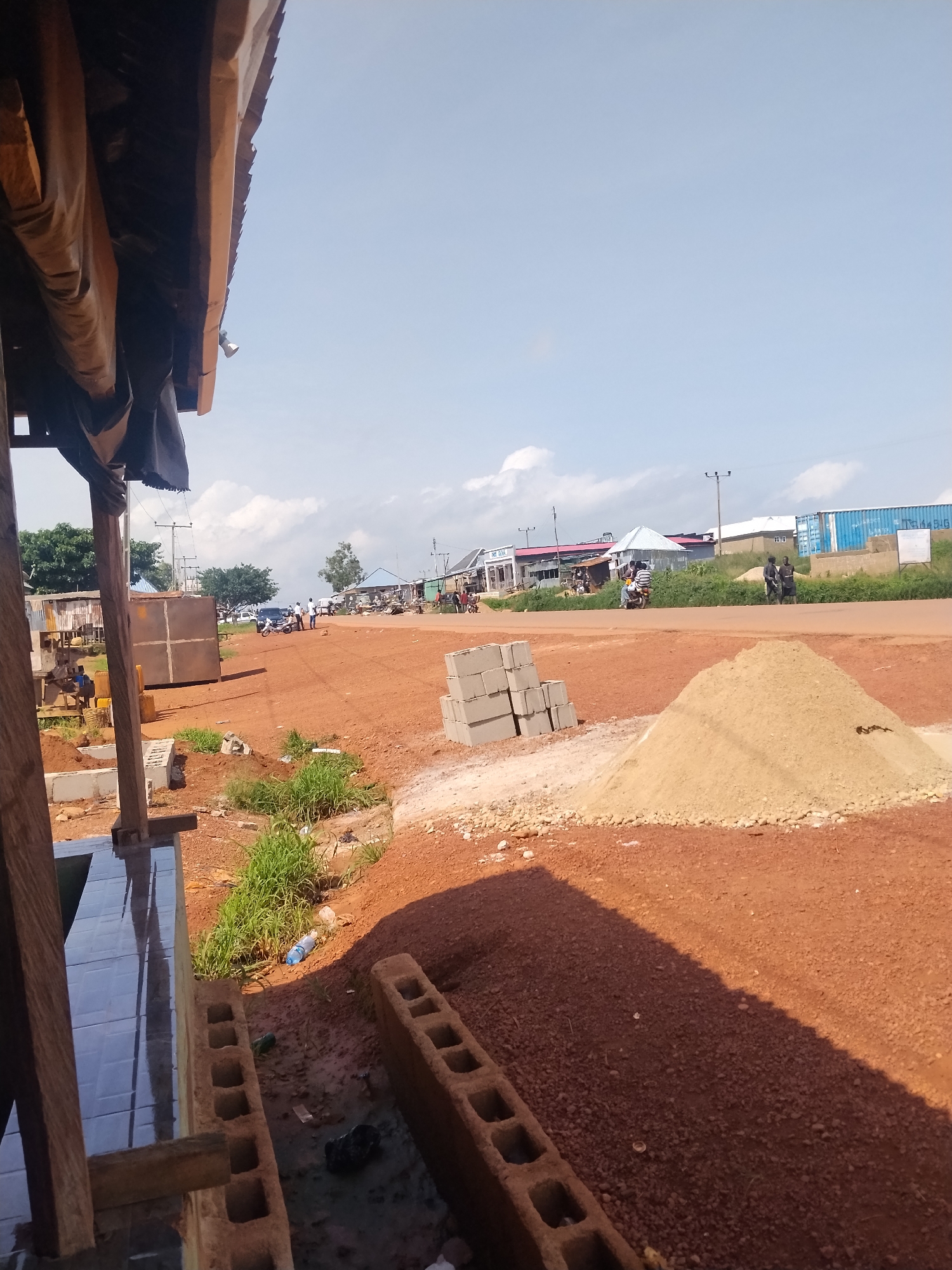 The image depicts an outdoor scene on a clear, sunny day. In the foreground, unfinished construction is visible, including cinder blocks and a pile of sandy soil. A tarp is partially covering a wooden structure on the left. The middle ground shows a dirt road with several individuals and a motorcycle. In the background, there are buildings, a blue shipping container, and trees. The overall environment appears to be a semi-urban or developing area.