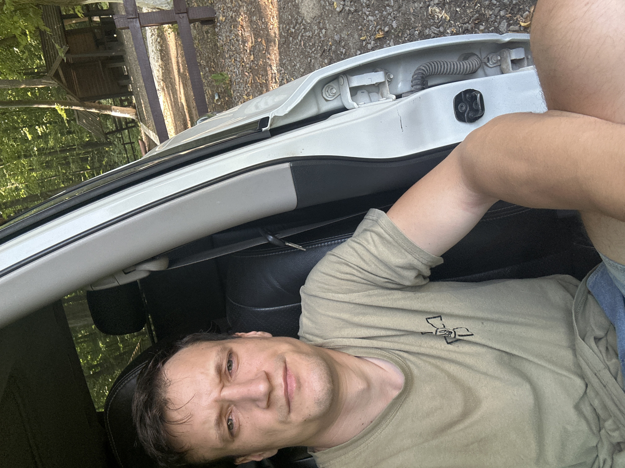 A man sits in the driver's seat of a white vehicle. He is wearing a light green t-shirt and blue shorts. The car door is open, revealing the interior and exterior of the vehicle. In the background, a wooded area with a wooden structure and a fence can be seen. The image appears to be taken during the daytime, with sunlight filtering through the trees. The location is described as Shatava, Ukraine.