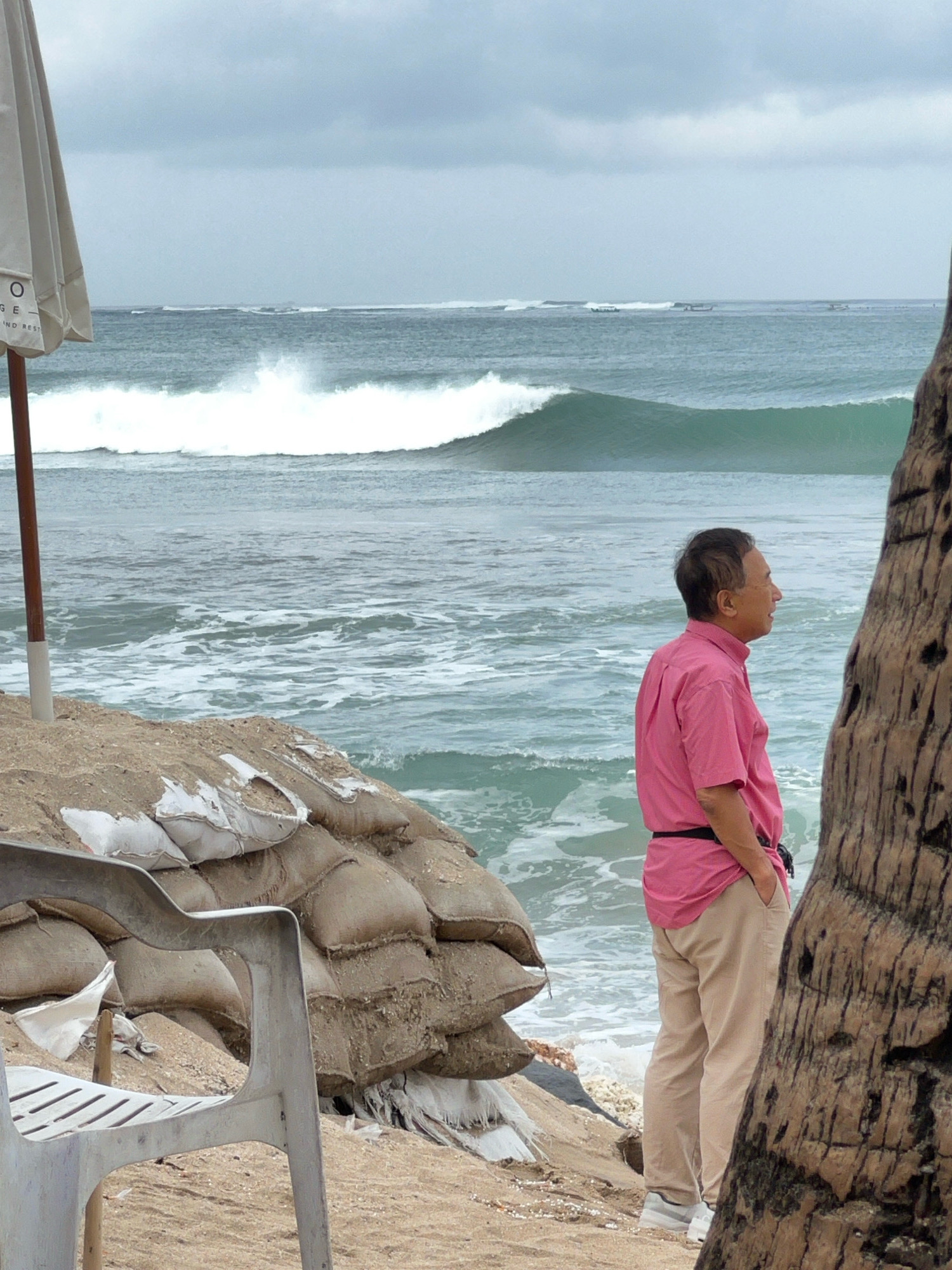 A man in a pink collared shirt and tan pants stands on a sandy beach, looking out at the ocean. He has his left hand in his pocket. Behind him, a large pile of sandbags is stacked, likely to prevent erosion. To his left, a portion of a white beach umbrella pole is visible. The ocean has large waves breaking, and the sky is overcast. The scene appears to be in Kuta, Indonesia, a popular tourist destination known for its beaches and surf. A large palm tree trunk is in the foreground on the right side of the image. A plastic beach chair is visible in the lower left corner.