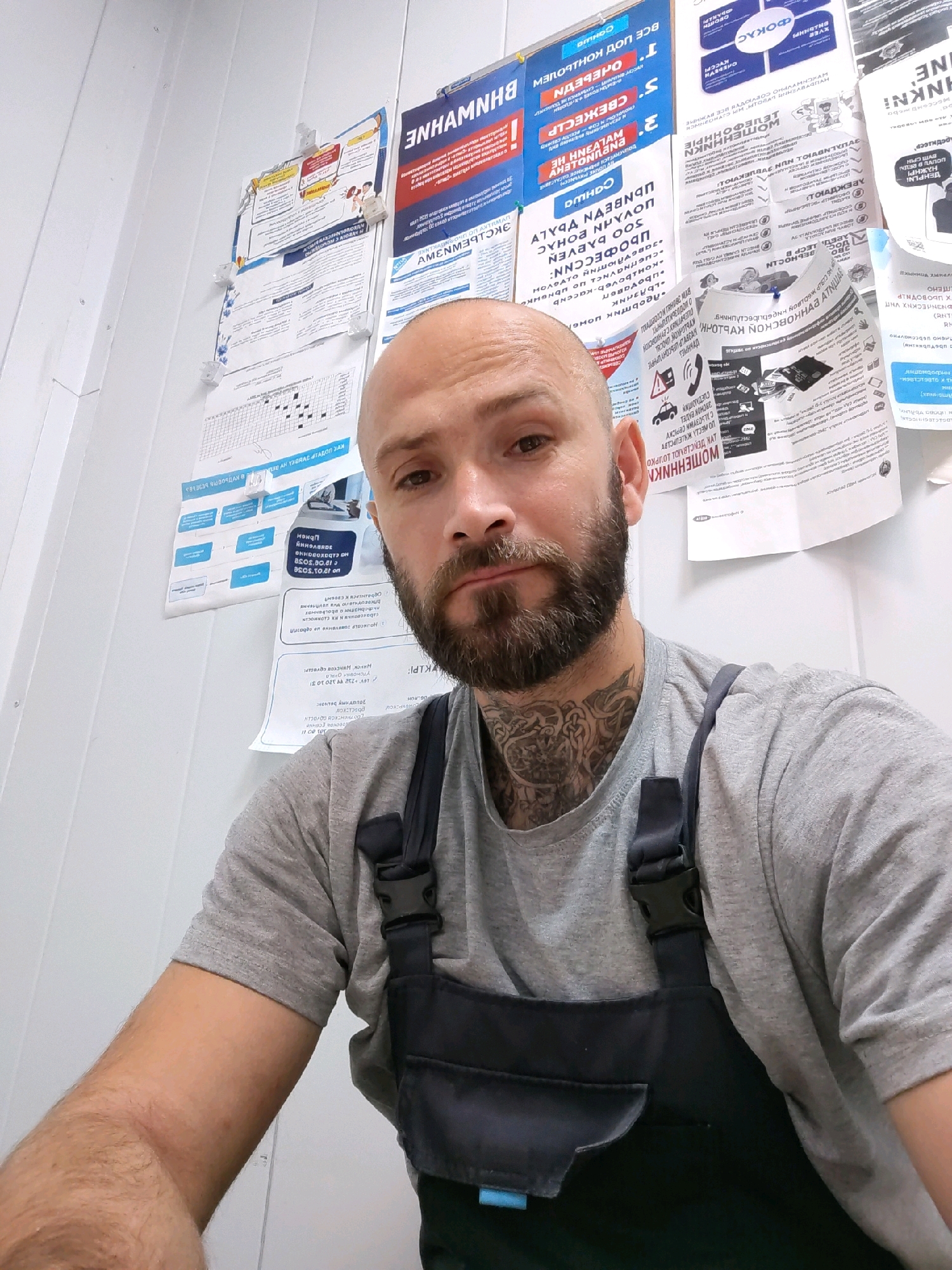 The image shows a close-up, selfie-style portrait of a bald, fair-skinned man with a beard and a tattooed neck. He is wearing a grey t-shirt and dark blue overalls. He is looking directly at the camera with a neutral expression. The background consists of a white wall covered with various posters and papers, some of which are written in Cyrillic script. The lighting appears to be artificial, coming from overhead. The overall scene suggests an indoor environment, possibly a workshop or office. The man's clothing and the posters in the background may indicate he is in a professional setting. Without further information, it is not possible to determine the specific location within the city of Pinsk, Belarus, but the presence of Cyrillic script suggests a Slavic-speaking region.