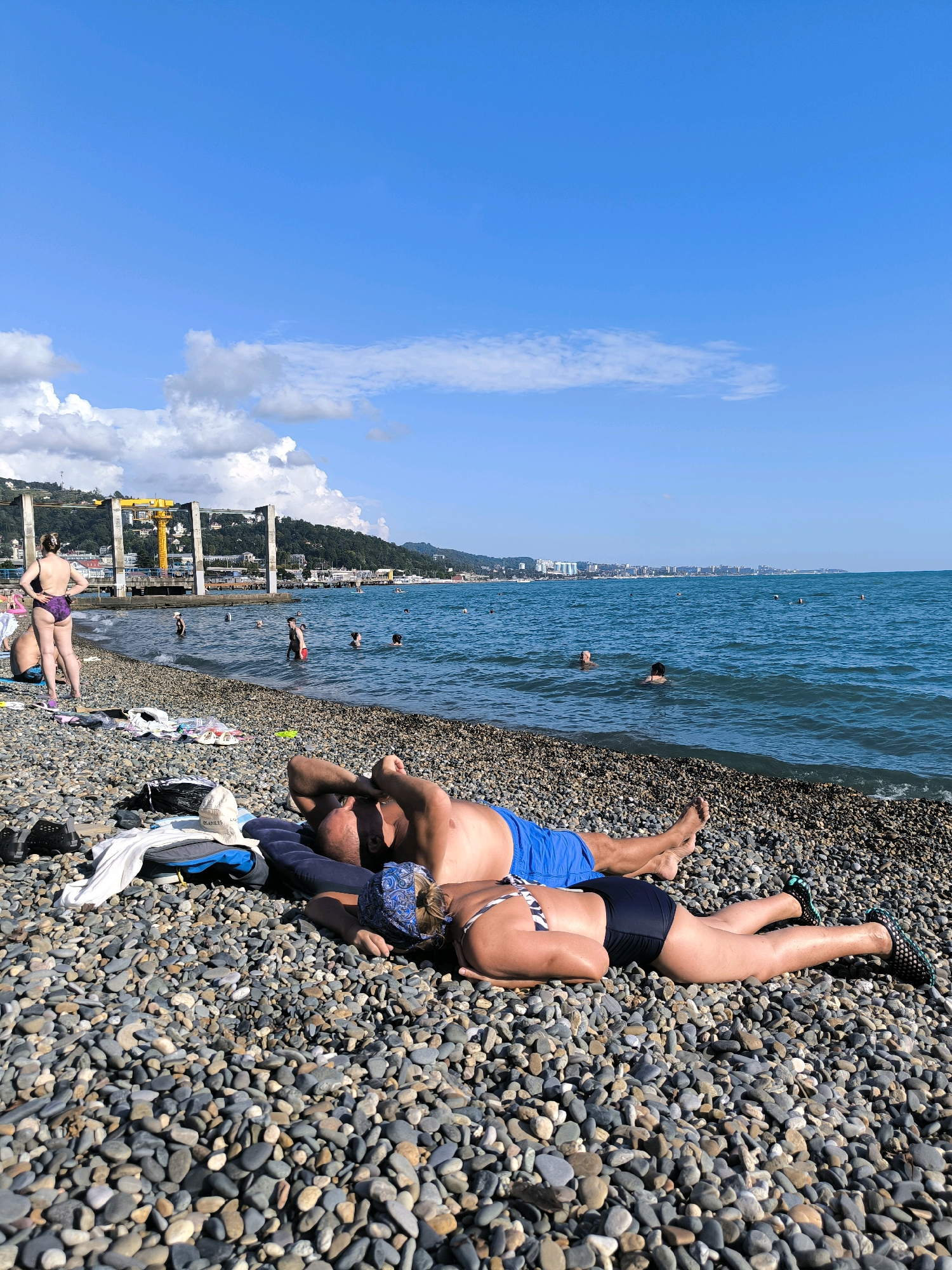A wide-angle, eye-level shot captures a pebble beach in Sochi, Russia, on a bright, sunny day with a clear blue sky and scattered clouds. In the foreground, two individuals are lying down on a blue inflatable mat, facing away from the camera. The male, on the left, is wearing blue swim trunks and has his arms behind his head. The female, on the right, is wearing a black bikini and a blue patterned bandana. Several items of clothing and accessories are scattered around them on the pebbles, including a white t-shirt, a black bag, and a beige hat.
The middle ground of the image shows the turquoise sea with several people swimming and wading. The shoreline is a mix of pebbles and water. In the background, a long stretch of coastline is visible, with buildings, hills, and a large yellow construction crane situated on a pier. The overall scene depicts a typical summer day at a beach resort.