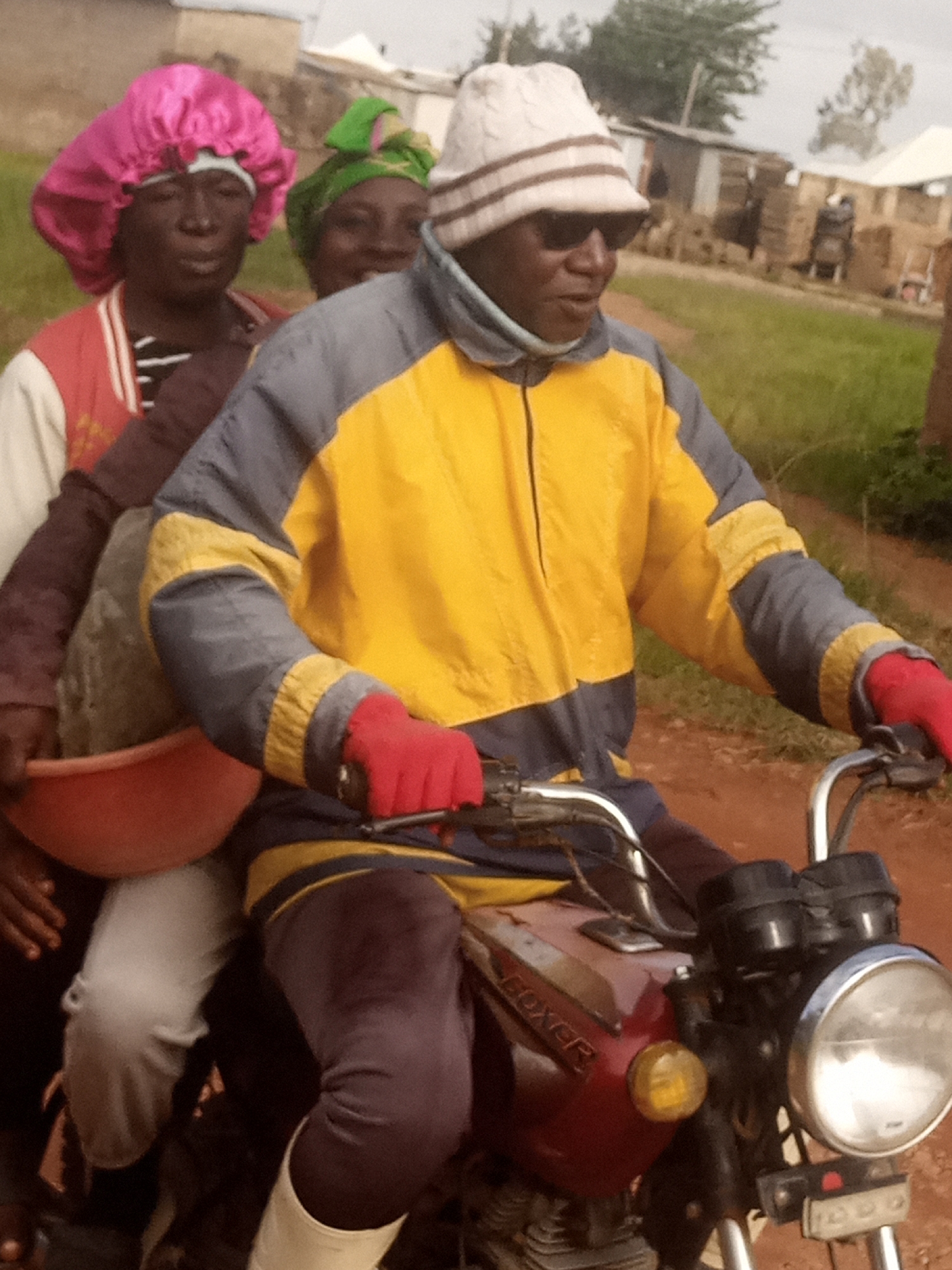 In the city of DU, Nigeria, three individuals are seated on a motorcycle, which is on a dirt road. The person in the front is wearing a yellow and grey jacket, red gloves, dark sunglasses, and a knitted hat. They are holding the handlebars of the motorcycle. Behind them, a person in a pink bonnet is also on the motorcycle, holding a red bowl with a rock in it. Another person, wearing a green headscarf, is positioned behind the person in the pink bonnet. The environment appears to be rural, with grassy areas and some buildings in the background.