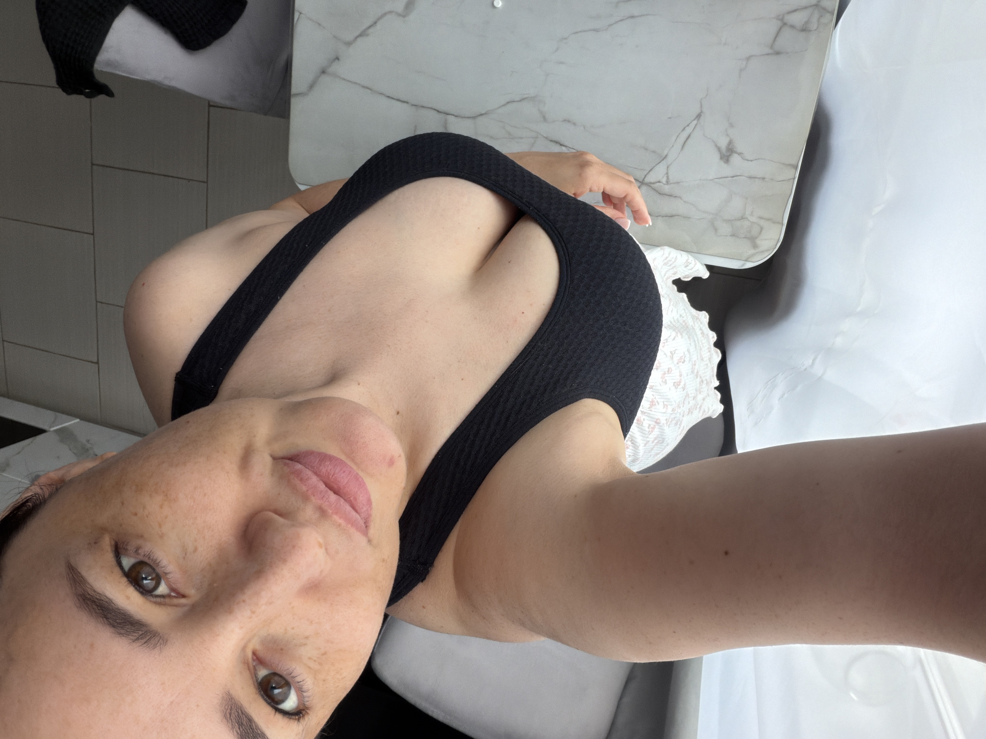 A high-angle, indoor selfie captures a woman in Водоканал Снт, Russia. She has fair skin with prominent freckles across her nose and cheeks, and dark brown eyes looking directly at the camera. Her dark hair is pulled back, and her lips are closed, tinted with light pink lipstick. She wears a black, textured, low-cut sports bra or tank top, revealing significant cleavage, paired with a white patterned garment with a ruffled edge, possibly a skirt or shorts. Her right hand (viewer's left) rests on a light gray and white marble-patterned surface, which appears to be a table or countertop. The background features light gray tiled flooring, a light gray chair or sofa on the left, and another light gray chair on the right draped with a black woven item. The scene is brightly lit.