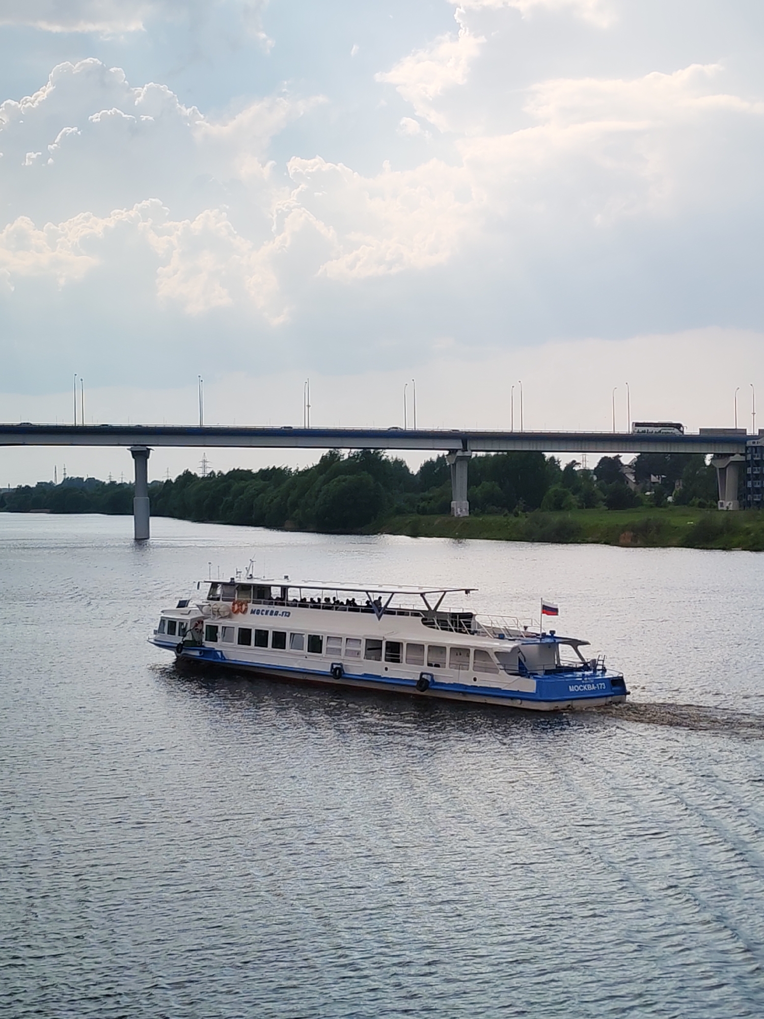 The image captures a passenger ferry boat, labeled "MOCKBA-173", navigating a river under a long, low bridge that appears to carry vehicular traffic. The boat is predominantly white with a blue trim, and has people visible on its upper deck. A Russian flag is attached to the stern. The water's surface is rippled, indicating movement of the boat and some current. The bridge stretches across the scene, supported by concrete pillars, with some vehicles visible on it. The background consists of a green tree line, suggesting the river flows through a wooded area, and several buildings. The sky above is cloudy, with light breaking through, which illuminates parts of the scene. The city is unknown.