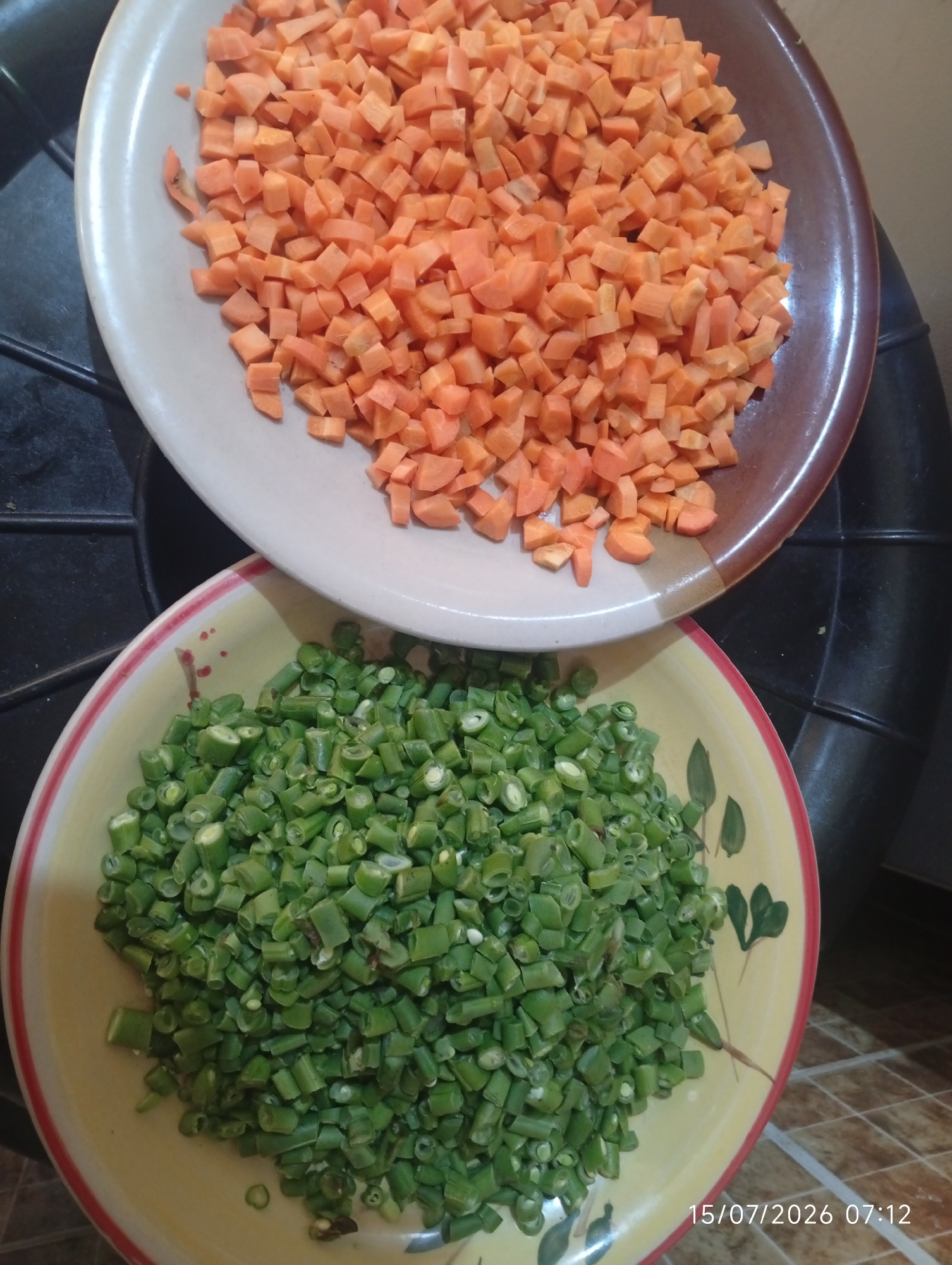 Two plates of chopped vegetables are displayed. The top plate, which is white with a brown rim, contains diced carrots. The bottom plate, a yellow bowl with a red and white rim and a floral design, is filled with chopped green beans. Both plates are positioned on a dark, textured surface, possibly a stovetop. The background is blurred, suggesting a kitchen environment. The lighting appears to be from an overhead source. A timestamp in the lower right corner reads "15/07/2026 07:12". The location is not visible or discernible from the image.