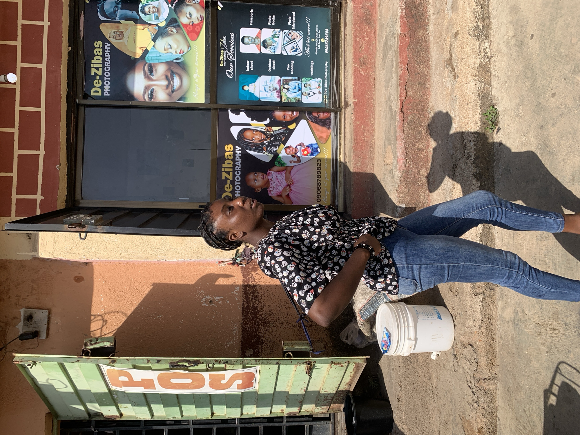 A photograph captures a woman outdoors, next to a building. She wears blue jeans and a black blouse with white skull motifs. A white bucket sits beside her. The building features a green awning with the word "POS" on it. Above the awning, there's a business advertisement for "De-Zibas PHOTOGRAPHY", along with a phone number and the phrase "Always at your service!!!". Further ad posters display various photography services, including "Indoor shoot", "Passports", "Outdoor shoot", "Photo Books", "Pre-wedding", "Photo Enlargement", and "weddings". The location is identified in the advertisement as "@Dudin Inuwa, COCIN Church, A.M Dung Street Jos", placing the scene in Jos, Nigeria.