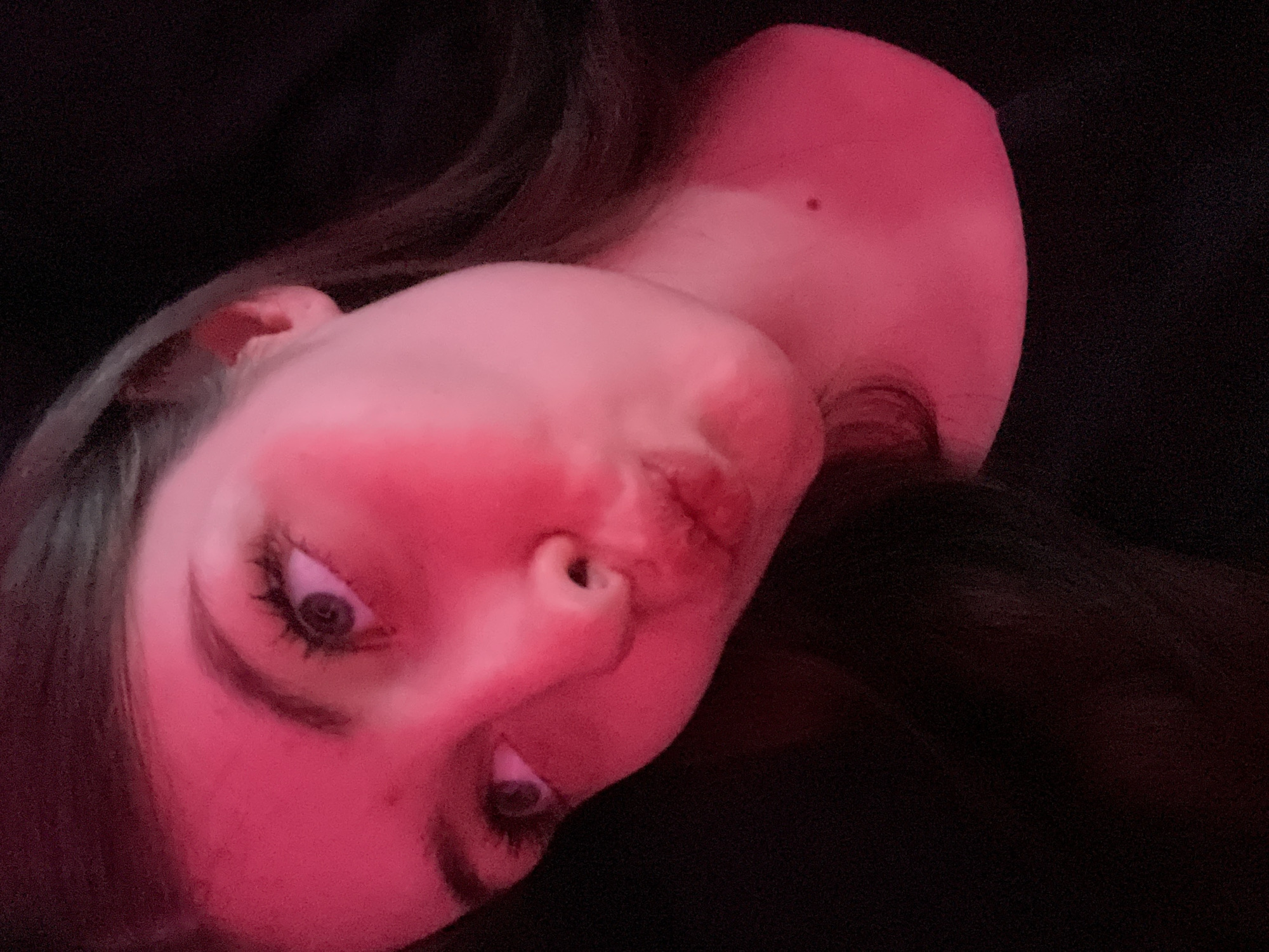 A close-up, slightly angled shot of a woman's face, predominantly lit with a red light source. The woman has fair skin, with visible eyes, nose, lips, and eyebrows. Her hair is dark and falls around her face. The background is dark and indistinct. The image doesn't show any specific location in Saint Petersburg, Russia, or any other discernible location. There are no visible actions, interactions, or other subjects/objects besides the woman's face.