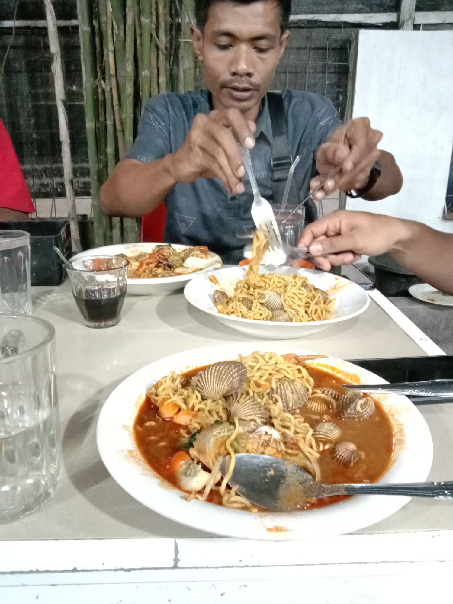 The image depicts a man dining at a table, likely in a restaurant, with two plates of food prominently displayed. The food appears to be a noodle dish, possibly Mee Goreng Aceh, with what seem to be cockles or similar shellfish. One plate has a brown, liquid-like sauce surrounding the noodles and cockles. Utensils, including a spoon and fork, are visible. The man is using a fork to eat. Another person's hand is visible in the image. There are glasses with liquid on the table. The background includes a bamboo-like structure and a white wall. The city of Lhokseumawe, Indonesia, is provided as the possible location.