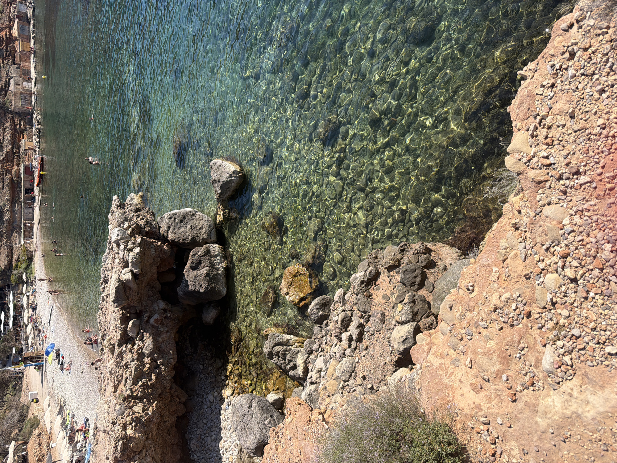 This media file depicts a picturesque coastal scene in Sant Antoni de Portmany, Spain, likely during a sunny day. The viewpoint is from a cliff overlooking a beach and the clear, shallow waters of the Mediterranean Sea. The water's transparency allows the viewer to see the rocky seabed below, illuminated by dappled sunlight.
On the beach, there are numerous beachgoers enjoying the day. Lounging chairs and umbrellas are scattered across the pebble and sand shore, indicating a popular tourist spot. Several people are seen swimming or wading in the sea, while others are relaxing on the beach. Some buildings, appearing to be small structures or beach huts, are nestled into the cliff face on the left side of the image.
The rocky cliff face itself is a prominent feature, with rough, reddish-brown rock and smaller stones. Patches of dry vegetation are visible, adding to the natural, rugged landscape. The overall impression is one of a vibrant, sunny day at a Mediterranean beach, with a mix of natural beauty and human activity. The clear water, the sunlight, and the relaxed atmosphere of the beachgoers suggest pleasant weather and a leisurely time. No visible text is present in the image.