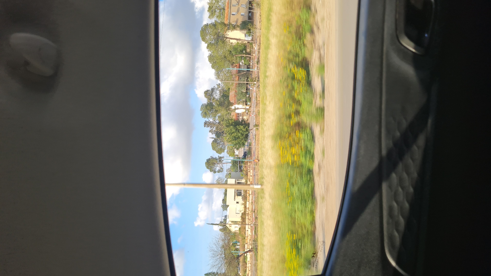 A roadside view from inside a moving vehicle in Bnei Shimon Regional Council, Israel. Outside, a grassy field with yellow wildflowers is adjacent to a residential area with multi-story buildings, trees, and utility poles under a blue and cloudy sky. The view is framed by the car's interior, including the headliner and door panel.