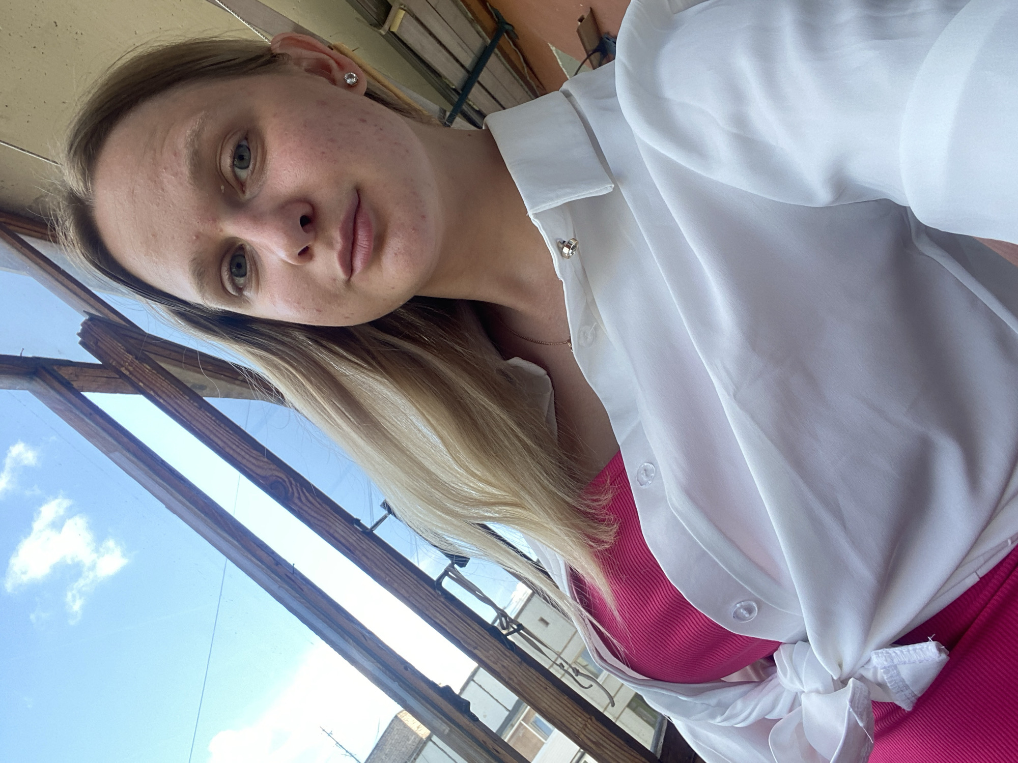 A young Caucasian woman with blonde hair and light blue eyes is taking a selfie. She is wearing a pink tank top and an unbuttoned white collared shirt. The woman has a stud earring in her right ear. The background appears to be outdoors, with a clear blue sky and some clouds visible through wooden beams. Buildings and structures can be seen in the distance. The lighting suggests it is daytime. The image quality is good, with no significant artifacts.
