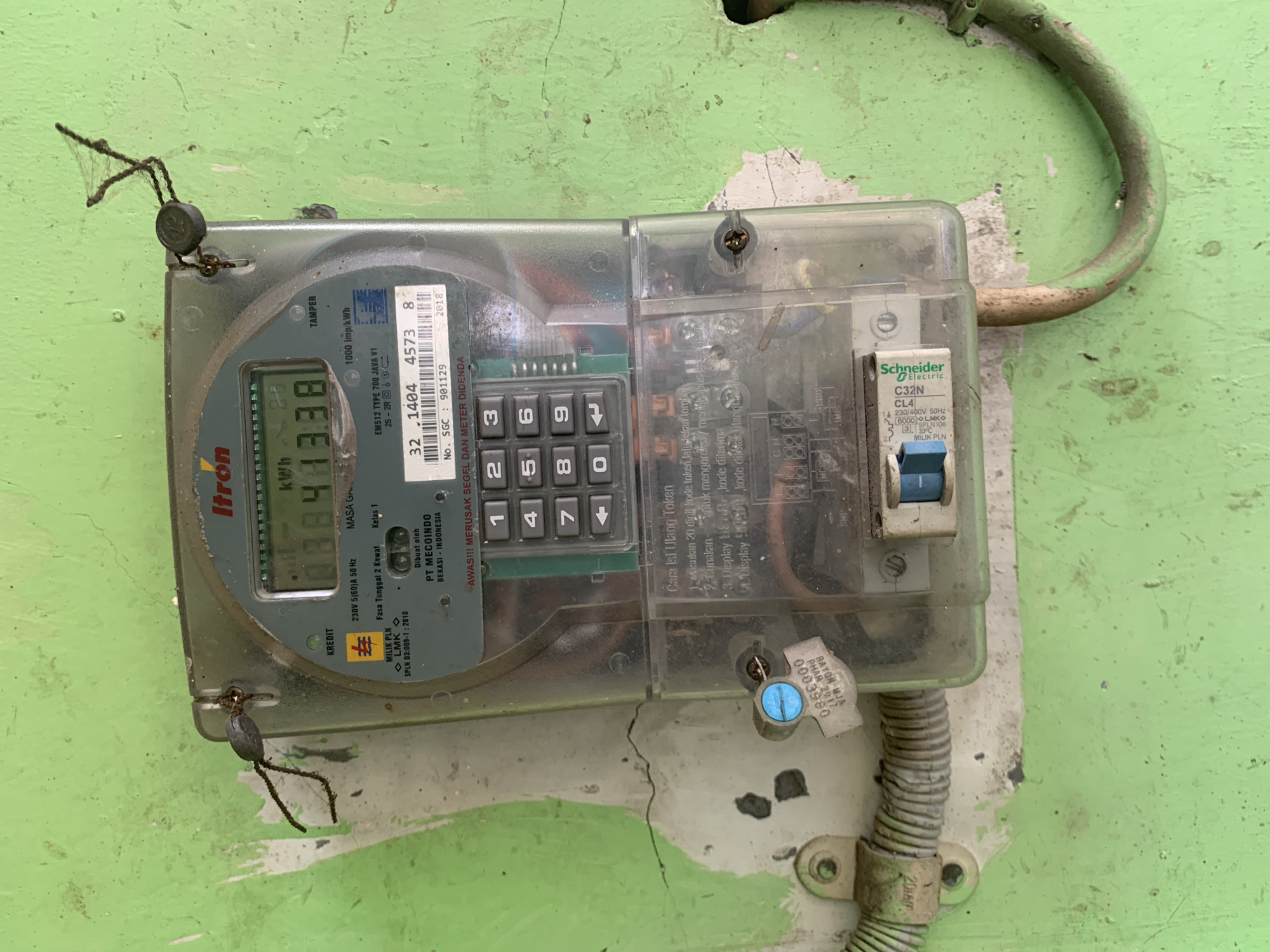 The image shows a close-up of an electricity meter mounted on a green, distressed wall in Dukuh Mojo, Indonesia. The meter, a clear plastic "Itron" brand digital display model, shows a reading of "088338" kWh. Below the digital display, a keypad with numbered buttons (0-9) and arrow keys is visible. The meter has various labels, including "KREDIT", "TAMPER", and details about its specifications. A small, blue-tagged seal is attached to a wire leading into the meter. To the right of the meter, a Schneider Electric circuit breaker labeled "C32N" is visible. The wall surrounding the meter shows signs of wear and tear, with peeling paint and cracks. There is also some evidence of organic matter, possibly cobwebs, attached to the top of the meter and on wires. The overall environment appears to be an outdoor or semi-outdoor setting, common in residential areas. No people are visible in the image.
