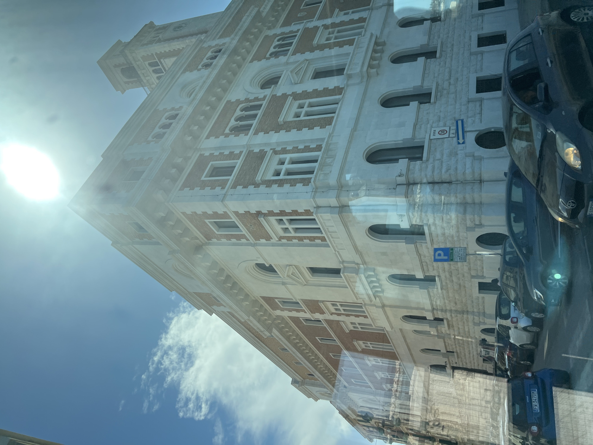 In Bari, Italy, a wide-angle, low-angle shot captures a sun-drenched building with classical architecture. The building features a prominent tower and is adorned with decorative brickwork and white stone accents. Sunlight glares intensely at the top of the frame. In the foreground, several cars are parked and driving on a street, their reflections visible on a glass surface, likely a window. A blue Smart car with license plate "FW 347DM" is partially visible on the left. A speed limit sign indicating "30" and a directional arrow are posted on the right side of the street. The sky above is a bright blue with scattered white clouds.