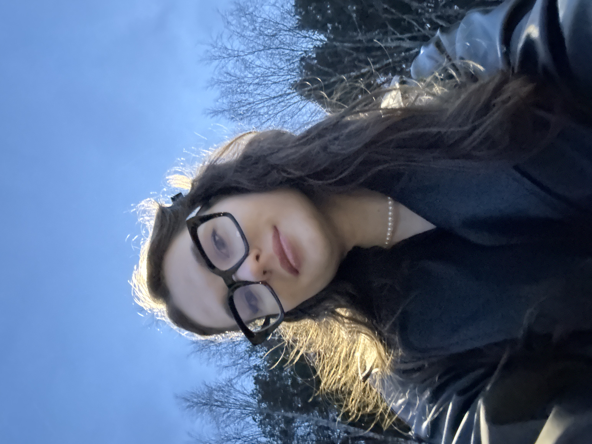 The image depicts a selfie taken outdoors in Chaikovsky, Russia. The subject is a young woman wearing black-framed glasses and a pearl necklace. She has long, dark hair and is wearing a dark-colored garment. Her tongue is slightly visible, indicating a playful expression. The background shows a blue sky with bare trees. The lighting is bright, suggesting daytime, with sunlight causing a halo effect around her head.