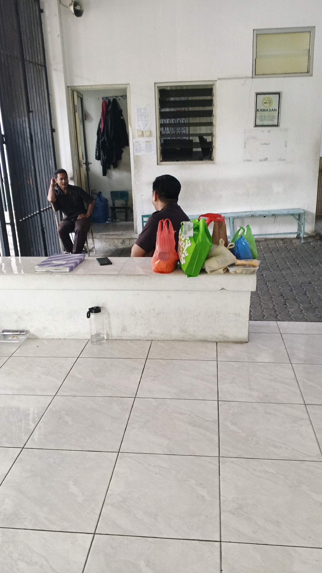 In Semarang, Indonesia, a scene unfolds in what appears to be a public waiting area or office. Two men are present. One man, wearing a dark brown uniform, is seated on a chair to the left, with his arm resting on his head as if in thought or weariness. He is positioned near a large metal gate. The other man, also in a dark uniform, sits on a bench on the right, facing away from the camera, with his back towards the first man. He is next to a white counter.
On the counter, a variety of colorful plastic shopping bags are placed: two orange, two green, one brown, and one blue. Beside these bags, there are some food containers and a book or folder. A black mobile phone is also visible on the counter. On the floor in front of the counter, a clear plastic water bottle with a black lid stands upright.
The environment features white tiled floors and walls. A doorway behind the man on the left reveals clothes hanging on hooks and large blue water dispensers. The wall on the right has windows with horizontal blinds and a framed sign that includes a smoking prohibition symbol and the word "KAWASAN." A long, light blue bench is positioned along the wall in the background. The overall impression is one of a somewhat utilitarian and orderly space, possibly a government building or a security checkpoint.