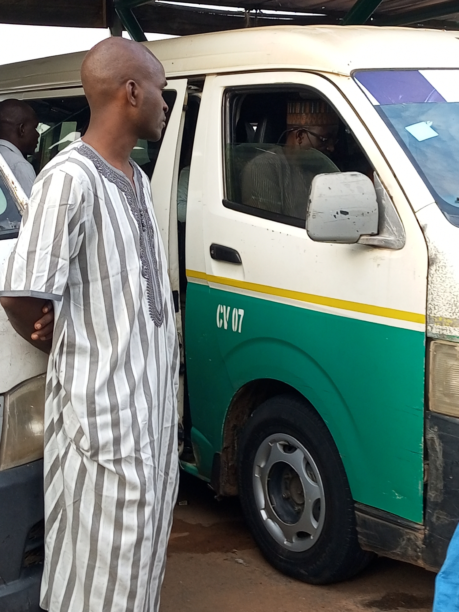 In Gombe, Nigeria, a bald man wearing a grey and white striped outfit stands beside an open door of a white van. The van is marked with "CV 07" in white lettering and has a green lower section with a yellow stripe separating the two colors. Inside the van, a man wearing glasses and a patterned hat is visible behind the steering wheel. The environment appears to be an outdoor setting with a dirt ground surface and what looks like a wooden shelter overhead.