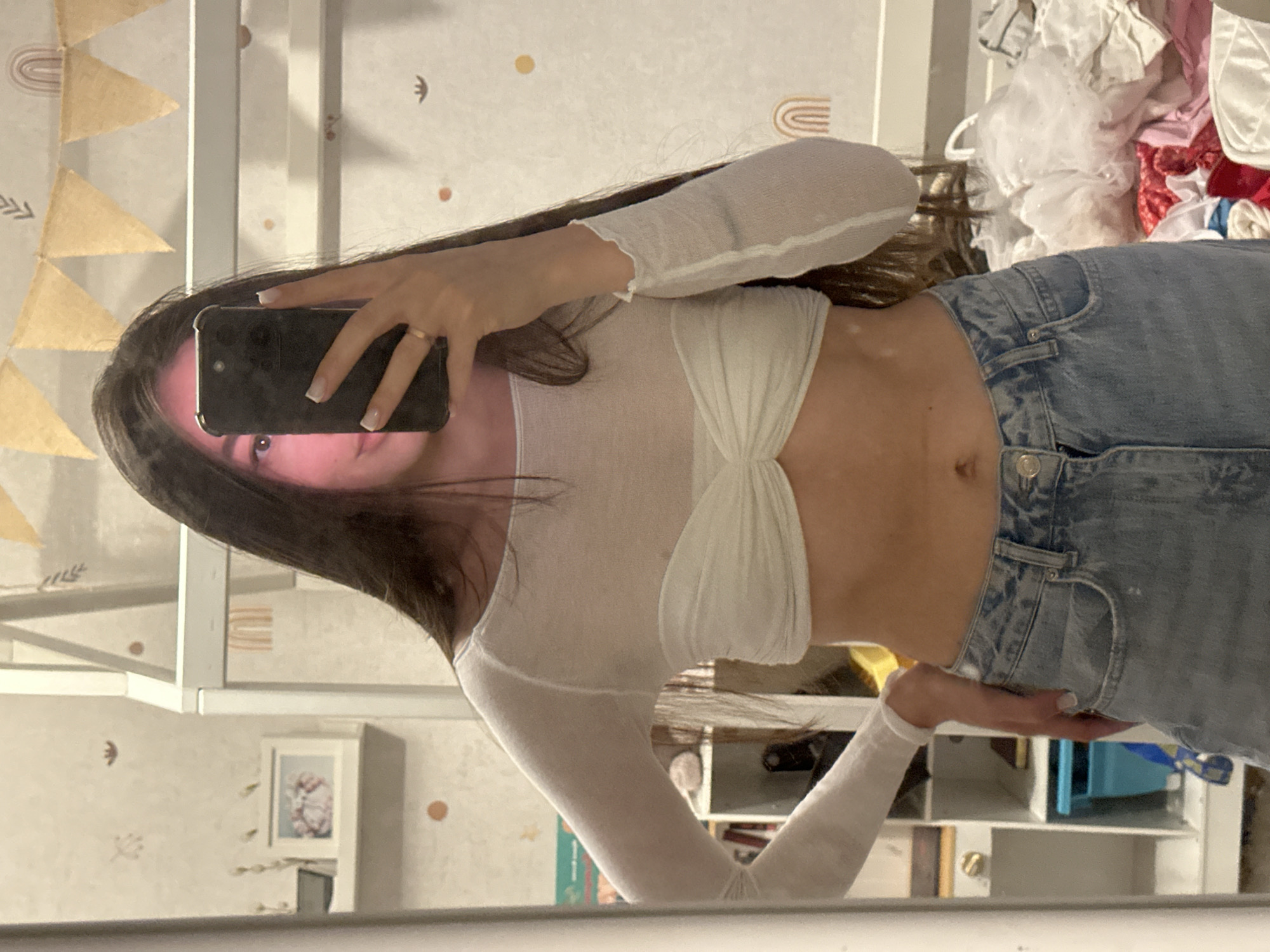 A woman takes a selfie in a mirror. She has long brown hair and is wearing a white long-sleeved top and blue jeans. She is holding a black smartphone to take the picture. The background shows a room with white furniture and decorations on the wall. No indication of Berezovka, Russia.