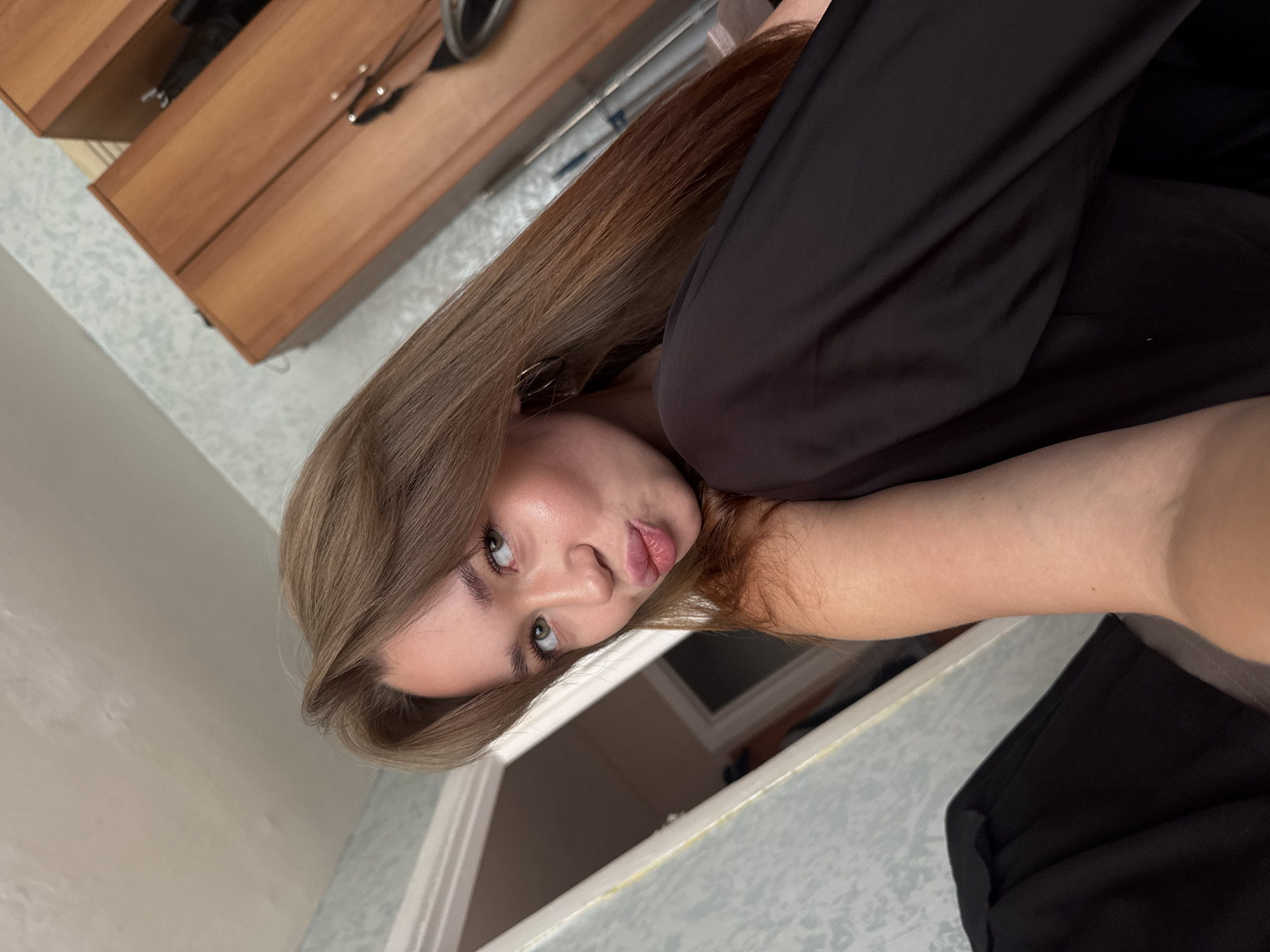 A young woman with shoulder-length brown hair and green eyes is shown in a close-up, angled shot. She is wearing a black top and has her hand on her arm, with her face turned towards the camera, puckering her lips. The background consists of a ceiling, a textured wall with a light blue and white pattern, and a wooden wardrobe. A doorway is visible to the left of the frame. The scene appears to be indoors, in what could be a bedroom or living space.