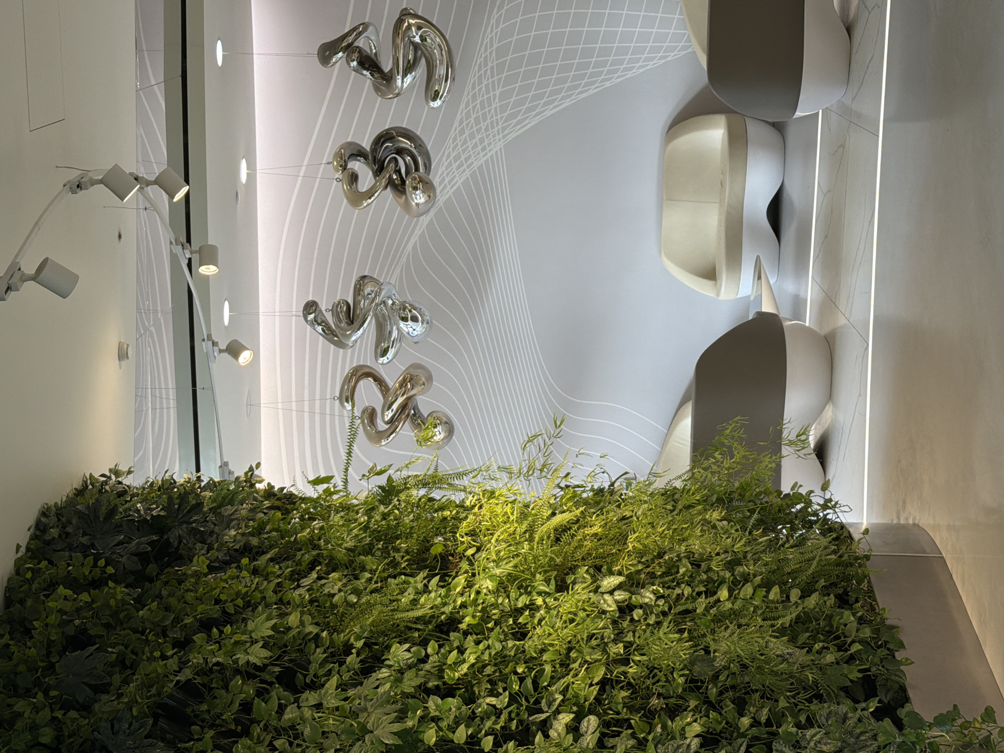 The image depicts an interior space, likely in Moscow, Russia. A vertical garden, comprised of various green plants, occupies the lower portion of the frame. Above the garden, silver-colored, abstract sculptures hang from the ceiling. To the right of the sculptures, modern seating arrangements, possibly chairs or benches, are visible. The background features a white wall with a subtle, flowing pattern. On the left, a wall is adorned with a series of modern, white spotlights.