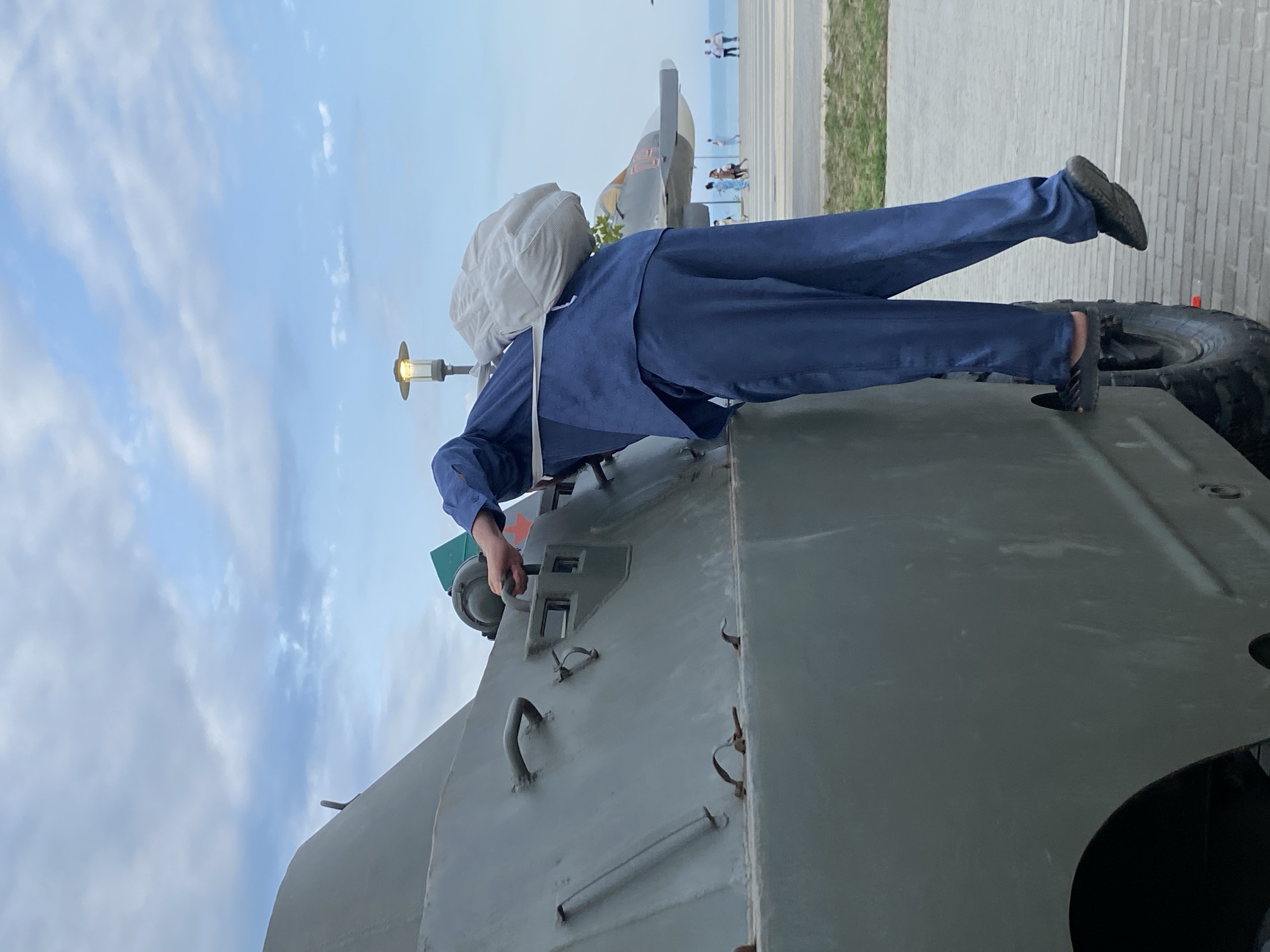 In Arablyar, Russia, a person in a blue jumpsuit and carrying a light-colored bag over their shoulder is climbing onto a green military vehicle. The person's left foot is on the vehicle's tire, and their right foot is on a step. The vehicle appears to be a BTR-60. In the background, there is a jet on display, a lamppost, a paved area, and a body of water. Several people are walking in the distance. The sky is cloudy.