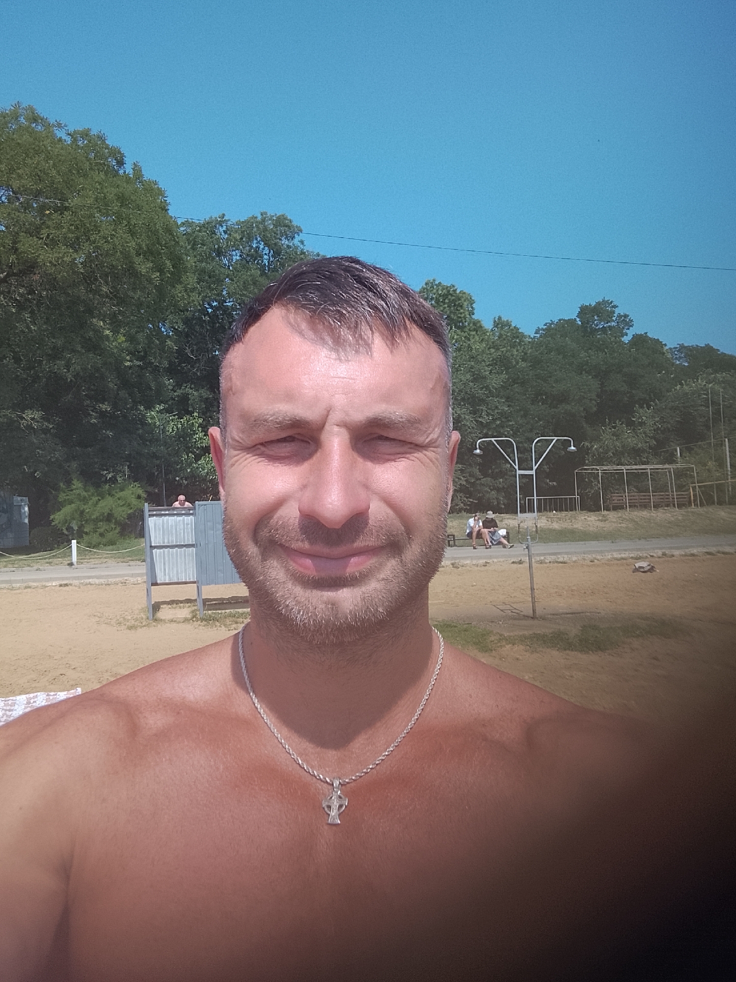 A middle-aged man with short brown hair and a beard is taking a selfie outdoors. He is shirtless, wearing a silver chain with a cross pendant. The background shows a sandy area, trees, and a clear blue sky. To the left of the man, there is a changing room structure, and further back, two people are sitting on a bench near an outdoor shower. The overall setting appears to be a beach or park area. The image does not contain any specific landmarks or visual cues that would allow for precise identification of the location within Chișinău, Moldova.