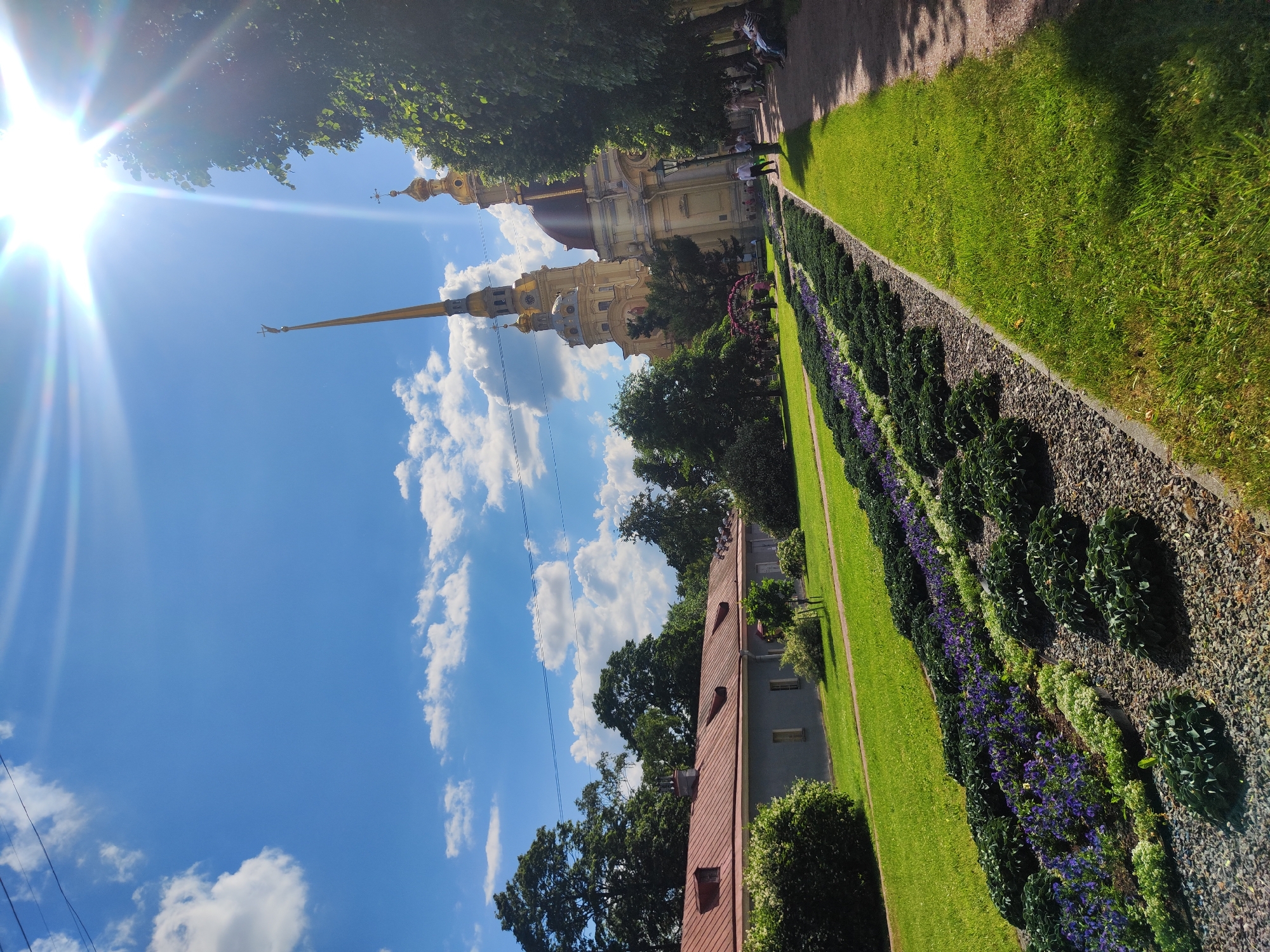 The image captures a sunny day at the Peter and Paul Fortress in Saint Petersburg, Russia. The Peter and Paul Cathedral, with its distinctive golden spire, dominates the background. In the foreground, a manicured garden features rows of purple and green flowers, bordered by a gravel path. A pathway lined with trees runs along the right side of the image, where several people are seen walking and sitting on benches. The sky is a vibrant blue, dotted with white clouds, and the sun is prominently visible, casting a bright glare. The fortress grounds appear well-maintained, with lush green lawns and mature trees.