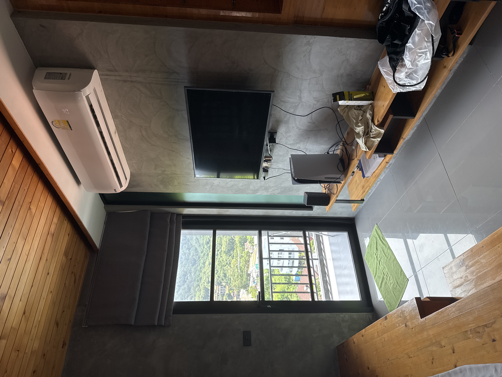 The image shows an interior view of a room in Rawai, Thailand, overlooking a lush, green hillside with scattered buildings. The room features a modern, minimalist aesthetic with concrete walls and a wooden ceiling. An air conditioning unit is mounted high on the wall. A large flat-screen television is centered on a concrete wall, mounted above a wooden shelf unit. On the shelf are a gaming console, a speaker, a rolled-up shirt, a yellow box, and a white plastic bag containing black items. A large glass door or window opens to a balcony with a railing, offering a view of the exterior landscape. A green welcome mat is placed on the grey tiled floor in front of the door. To the left of the door, a wooden bench or desk is visible. The scene appears to be a living space or bedroom within a residential building.