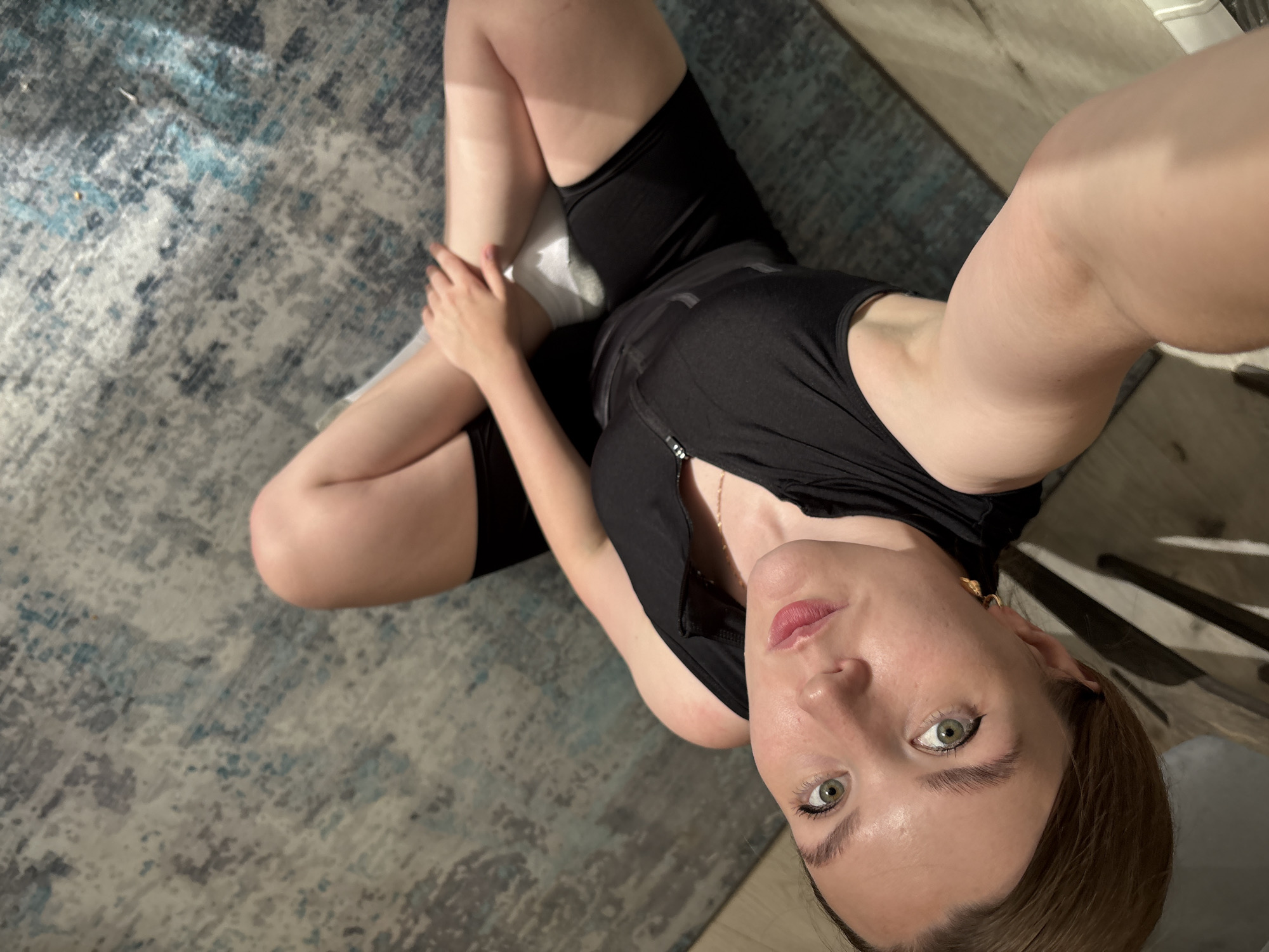 A young Caucasian woman with light skin and light brown hair is lying on her back on a patterned rug in a dimly lit room. She is wearing a black sports bra and black shorts. Her legs are bent and crossed at the knees, with her left leg resting on top of her right leg. Her left arm is bent at the elbow, with her hand resting on her right thigh. Her head is tilted back, and she is looking up at the camera. Her eyes are wide open, and her lips are slightly parted. The room appears to be a living room or bedroom, with a wooden floor and a rug. There is a wooden chair visible in the background. The lighting is low, with a spotlight from the right creating shadows on the woman's body. There is no visible indication of the city of Izhevsk, Russia.