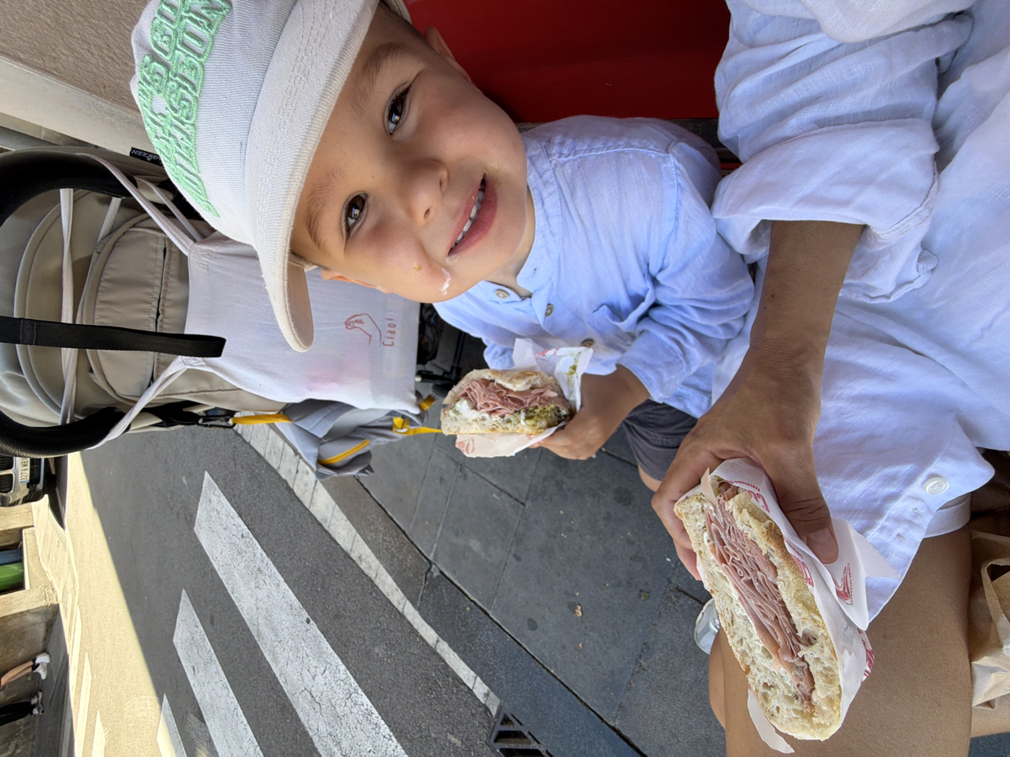 In Barcelona, Spain, a young boy wearing a white cap and a light blue collared shirt is holding a half-eaten sandwich. He has a smudge of white sauce on his cheek and is smiling. Nearby, an adult's hands are holding another sandwich. The scene takes place on a sidewalk next to a street with a crosswalk. A stroller and a car are visible in the background. The lighting suggests it is daytime.