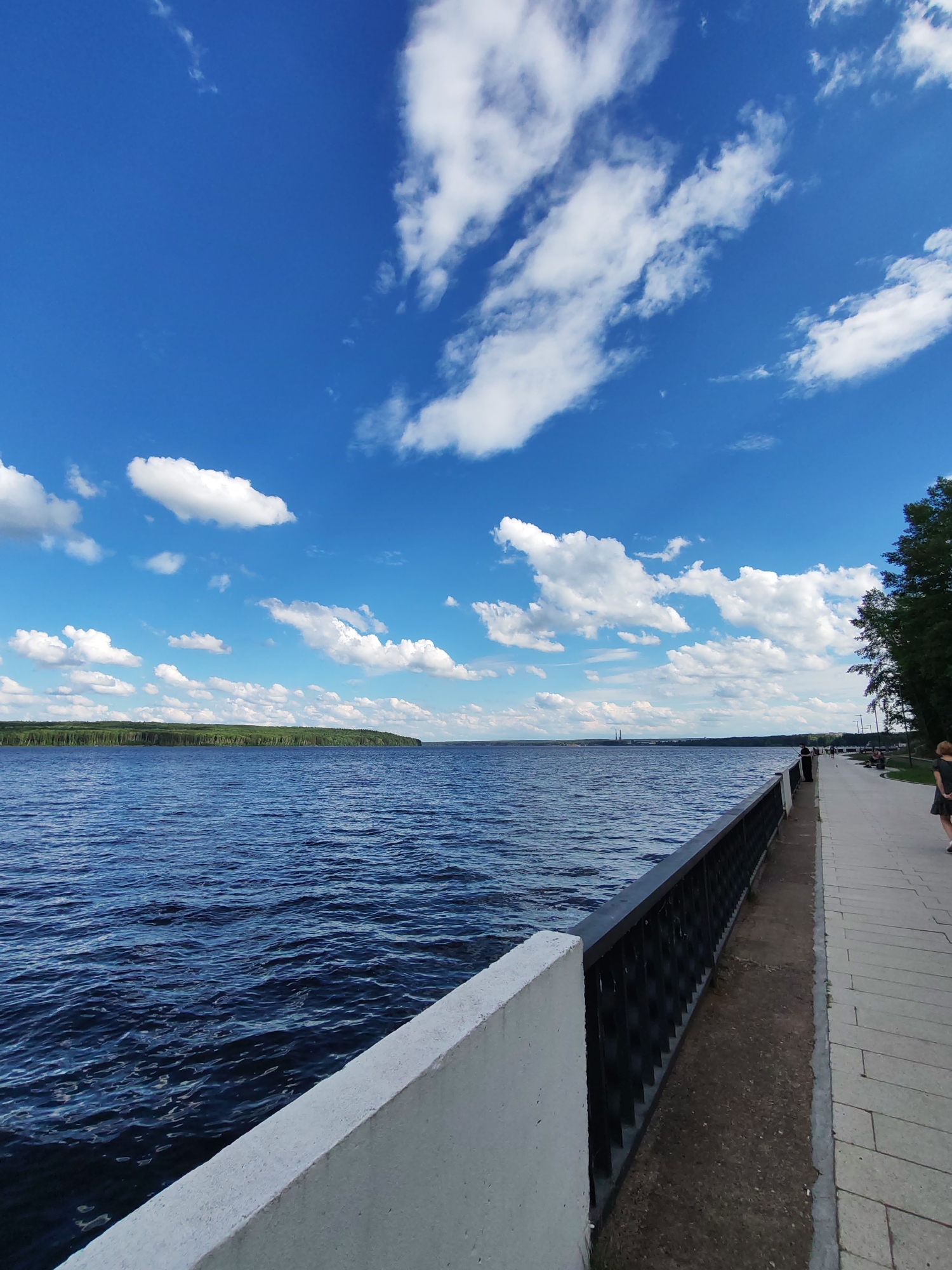 This is a photograph taken from a waterfront promenade in the city of Chaikovsky, Russia, facing a large body of water, likely the Kama River reservoir, with a distant tree-lined shore. The sky is a vibrant blue with scattered white cumulus clouds. A concrete retaining wall and a black metal railing run along the edge of the promenade, which is paved with stone slabs. Several people are visible on the promenade, walking and sitting on benches. One person, a woman with blonde hair, is walking away from the camera on the right side of the frame. The water is deep blue with moderate ripples, indicating a slight breeze. Trees line the right side of the promenade, casting shadows. The overall atmosphere is that of a pleasant day in a recreational area.