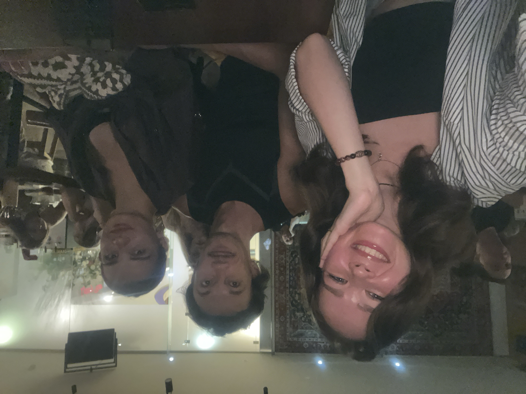 Three people are shown in the image, two men and one woman. They are possibly inside an establishment. The woman on the right is smiling, and she has her hand on her face. She is wearing a dark tank top and a striped button-down shirt. The man in the middle is wearing a black shirt. The man on the left is wearing a dark top, and there is a patterned shawl-like object behind him. There are other people seated at a table in the background. The room has a glass divider and a patterned rug on the floor. There are lights on the floor. The city of Nha Trang Ward, Vietnam, is mentioned, but there is nothing in the image that provides direct proof of this location.