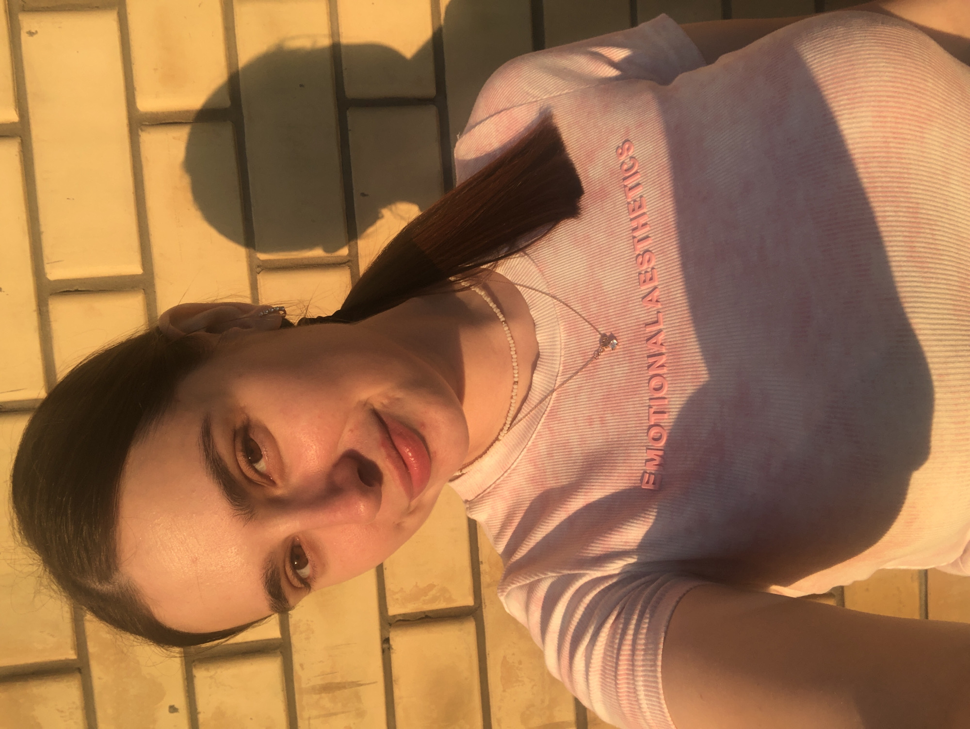 A young woman with dark hair pulled back in a ponytail is taking a selfie in front of a yellow brick wall. She is wearing a pink tie-dye t-shirt with the words "EMOTIONAL AESTHETICS" printed on it in pink lettering. A delicate beaded necklace with a small heart pendant is visible around her neck. The lighting suggests it is late afternoon or early evening, with long shadows cast by the woman on the wall behind her. The image was taken in Noviy, Russia.