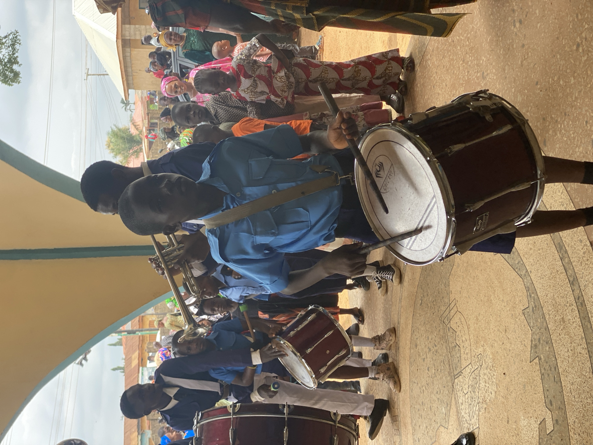 The image shows a marching band performing in what appears to be Kafanchan, Nigeria. The band members are wearing blue uniforms, and they are playing various instruments, including drums and trumpets. A crowd of people is watching the band perform. Some people are dressed in patterned cloth. The band and spectators are standing on a mottled tile ground under a covered pavilion. A building is visible in the background.