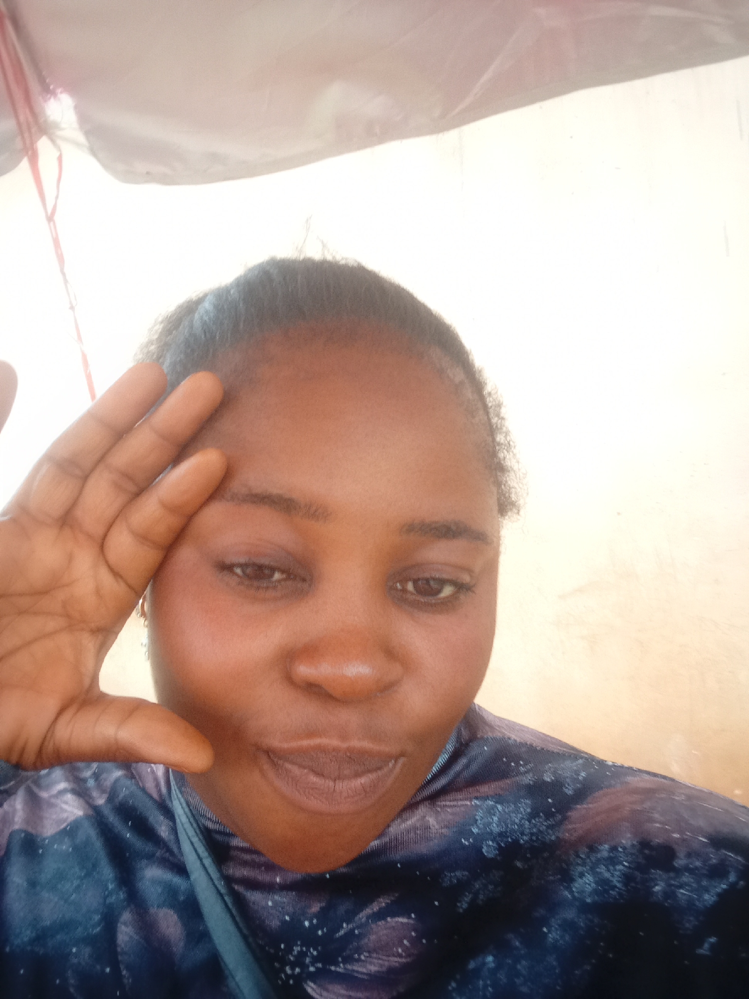 A woman of African descent is captured in a close-up, selfie-style portrait, her right hand raised to her forehead. She is wearing a dark, patterned top. The background appears to be an indoor setting with a light-colored wall and a hint of a white, possibly fabric-covered structure above. The lighting is natural and somewhat diffused. The image does not contain any visual elements that indicate its specific location within the city of Jos, Nigeria.