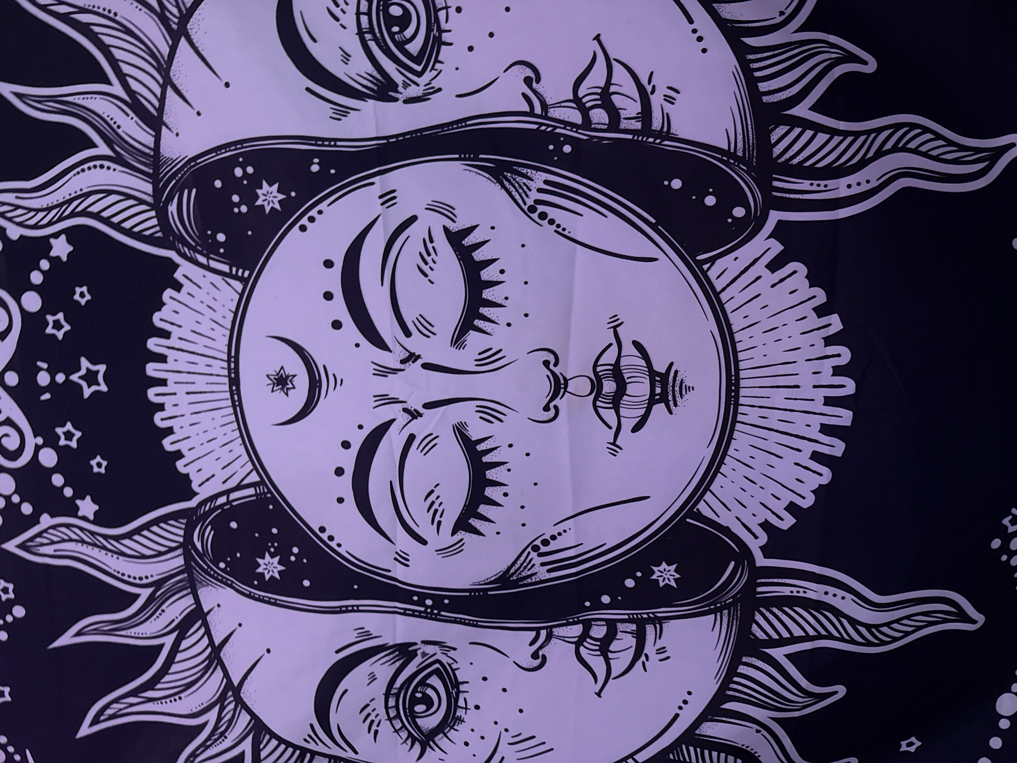 This is a close-up, head-on, medium shot of a black and white tapestry depicting three faces. The central face is a sun with closed eyes, a crescent moon on its forehead, and rays emanating from its head. Flanking the sun are two moon faces with open eyes. The left moon face is in profile, looking left, with eyelashes, freckles, and stylized lips. The right moon face is also in profile, looking right, with similar facial features. All three faces have dark outlines and are filled with black and white line work and dots, giving them a celestial, stylized appearance. The background is black and decorated with white stars and dots. The tapestry is lit with a purple light, giving it a lavender hue.