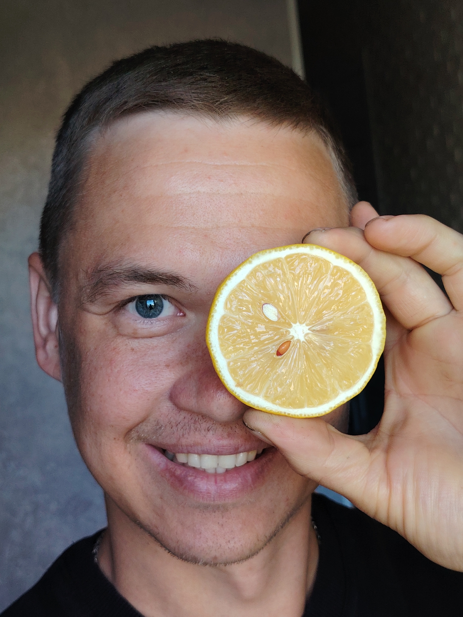 A close-up shot shows a fair-skinned man smiling with a slice of lemon held in front of one eye. The lemon is a cross-section with several seeds visible. The man has short, light brown hair, blue eyes, and some light stubble. He is wearing a black shirt. The background is blurry. The location is possibly Chaikovsky, Russia, based on metadata.