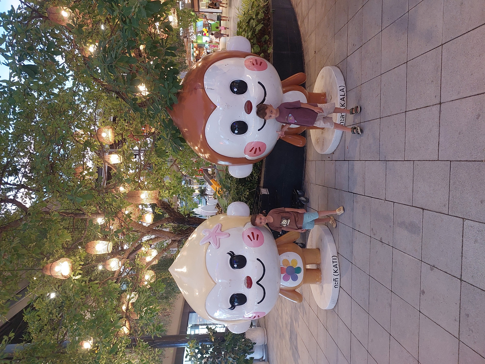 Two children pose with two large monkey statues in what appears to be an outdoor shopping area or walkway. The statues are cartoonish and brightly colored, with large heads and simple facial features. The statue on the left is brown and white with pink cheeks, and a child in a purple shirt and striped shorts stands next to it. The statue on the right is cream and blonde, adorned with a pink starfish on its head and a flower on its chest, and a child in a brown t-shirt and blue shorts stands beside it. Both children are smiling and appear to be enjoying themselves.
The background shows a lush green tree with numerous decorative lights hanging from its branches, creating a warm and inviting ambiance. In the distance, hints of shops and other patrons can be seen. The ground is paved with large, gray square tiles. The lighting suggests it is likely dusk or evening, as the artificial lights are prominent and the sky is dim.
Visible text on the base of the right statue reads "กะทิ (KATI)". The overall scene conveys a sense of fun and leisure, possibly during a vacation or outing.