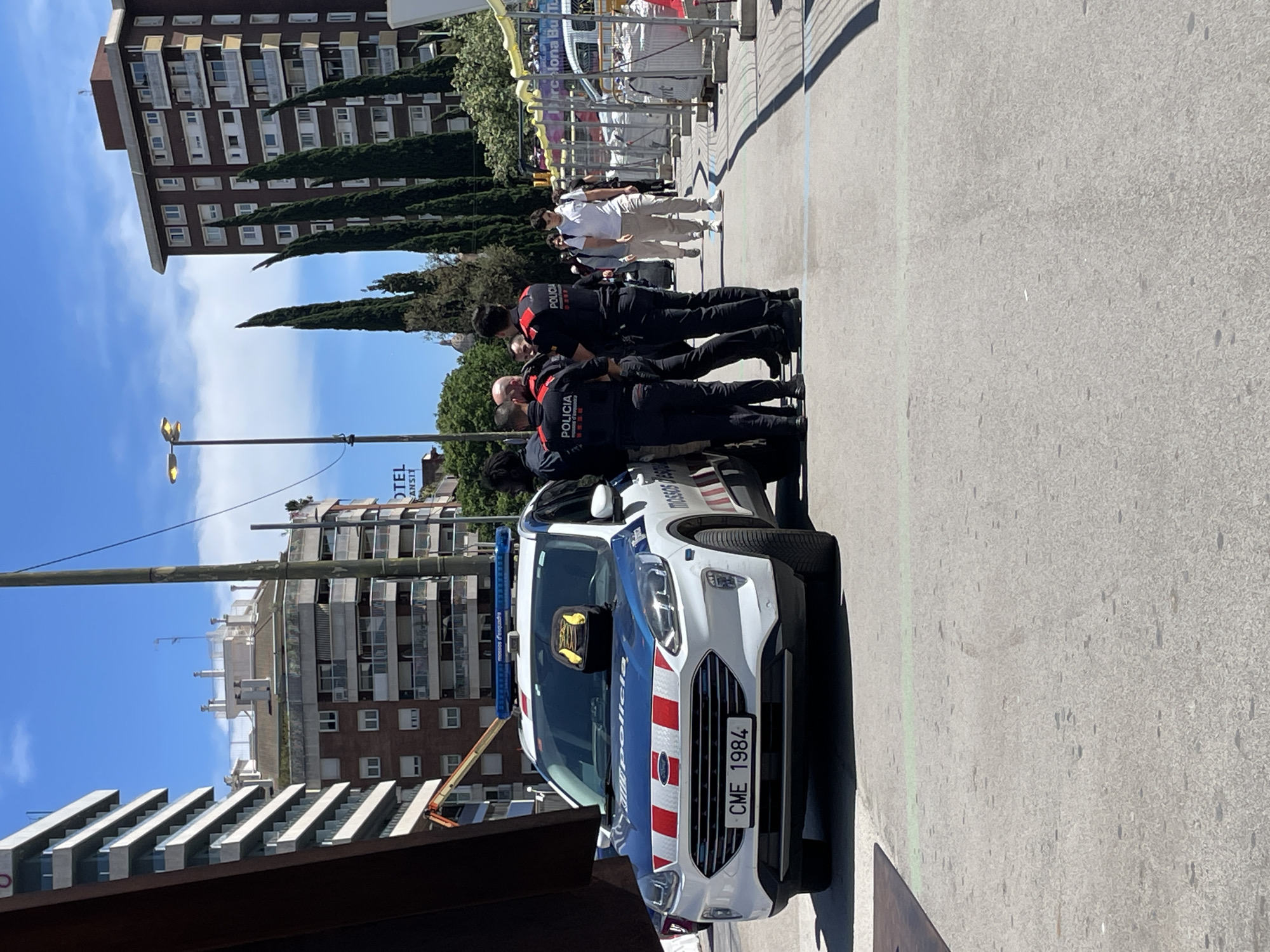 The image shows a police vehicle parked on a paved surface in Barcelona, Spain. The vehicle is a white and blue patrol car with red and white stripes on the front. The license plate reads "CME 1984." Several police officers in dark uniforms stand beside the vehicle, appearing to be in conversation. The surrounding environment includes buildings, trees, and a blue sky. The presence of what appears to be an event suggests the possibility that the officers are there to support or secure it.