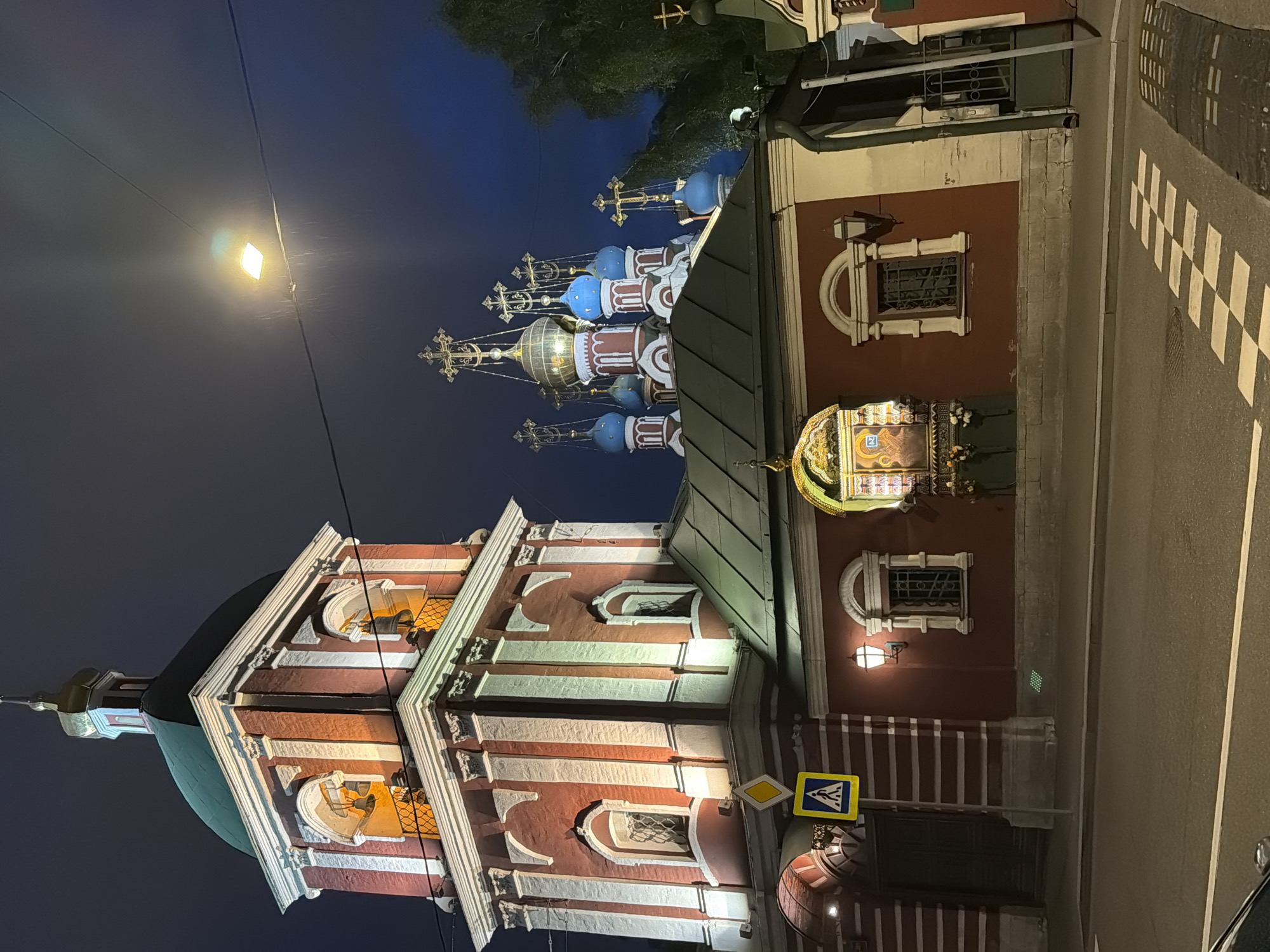 The image shows a building with a red and white facade, likely a church or religious structure, at night. The building has multiple domes with crosses on top. A street runs alongside the building, marked with a pedestrian crossing. Streetlights illuminate the scene. The location is possibly Перовское сельское поселение, Ukraine.