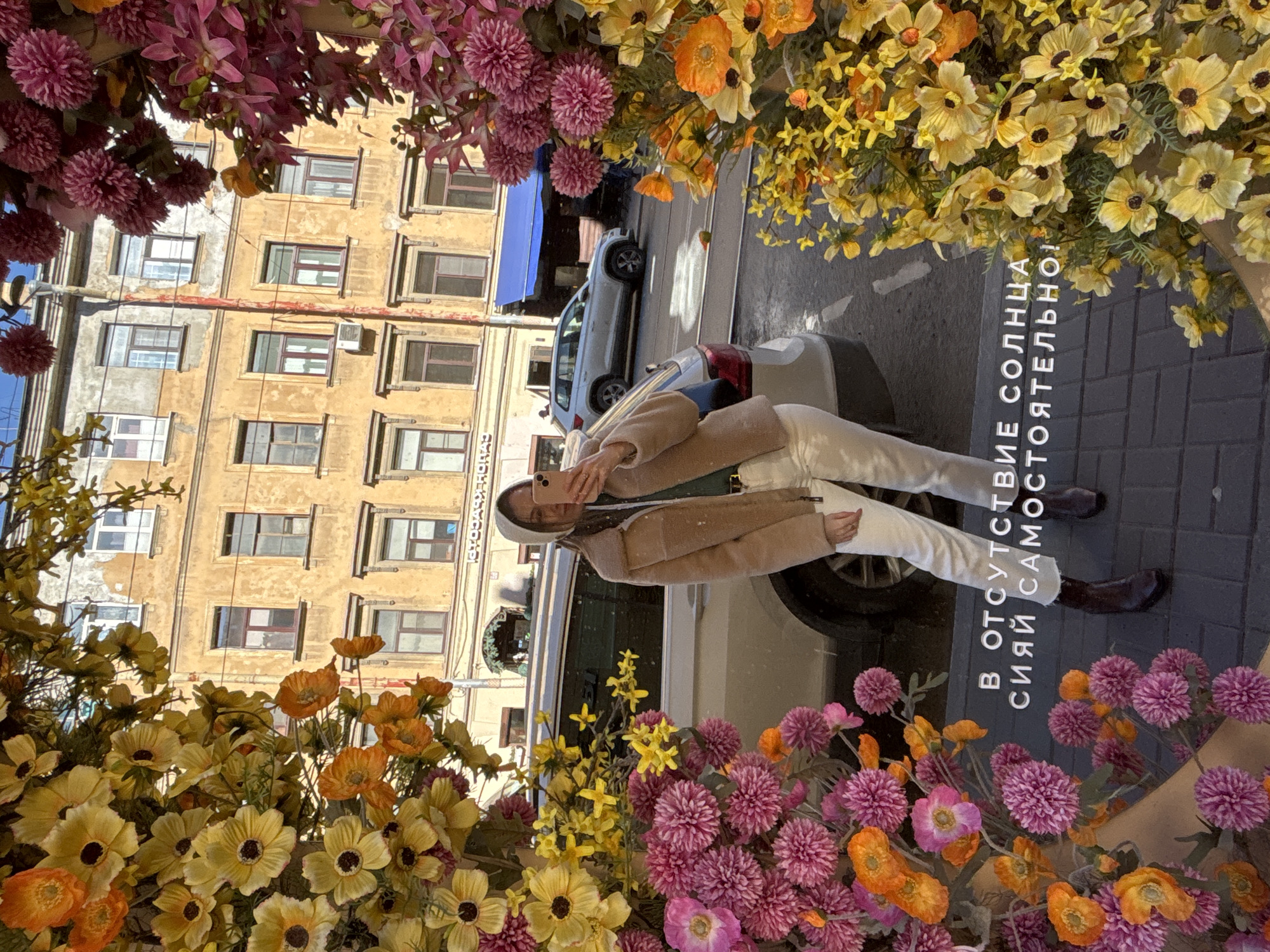 A medium shot captures a woman taking a selfie in front of a flower display, with a city street and building in the background. The image is framed by artificial flowers in various colors, including yellow, orange, and shades of pink and purple.
The woman is dressed in white pants and a light brown jacket. She is holding her phone up to take the picture.
Behind her, a silver car is parked on the street. The background features a multi-story building with rows of windows. The overall scene suggests a street in Saint Petersburg, Russia, given the architectural style of the building and the text in Russian language. The artificial flower display appears to be part of a shop or promotional setup along the sidewalk. The pavement is visible in the foreground. The image is rotated counter clockwise.