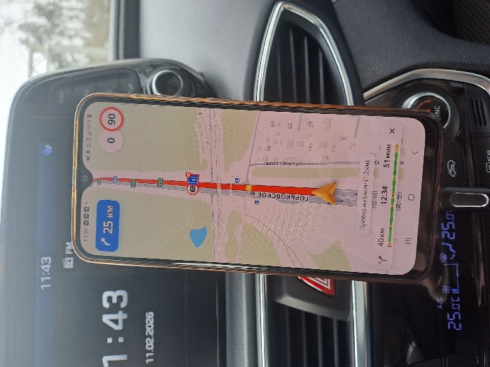The image depicts the interior of a vehicle, specifically focusing on a smartphone mounted vertically in a holder, displaying a navigation application. The scene is located in Obukhovo, Russia.
The smartphone screen shows a map centered on Gorkovskoye Shosse (ГОРЬКОВСКОЕ Ш.), with a yellow-orange arrow indicating the current vehicle position. A red line traces the route, and a traffic jam is indicated as "Пробка на 5 мин (1.2 км)" (Traffic jam for 5 min (1.2 km)). The navigation interface displays "25 KM" (likely distance to next turn or destination), "0 90" (possibly current speed and speed limit), total distance of "40 км," an estimated arrival time of "12:34," and remaining travel time of "51 МИН." The phone's status bar also shows "11:43."
In the background, the car's infotainment screen is visible, displaying "11:43" and the date "11.02.2026." A "FM" icon is also present. Below the smartphone, the vehicle's climate control panel shows a temperature setting of "25.0 °C" on both the left and right sides. A red hazard light button is partially visible to the left of the phone. The dashboard surfaces appear dark grey or black. The blurry background seen through the windshield suggests an outdoor environment with indistinct structures.