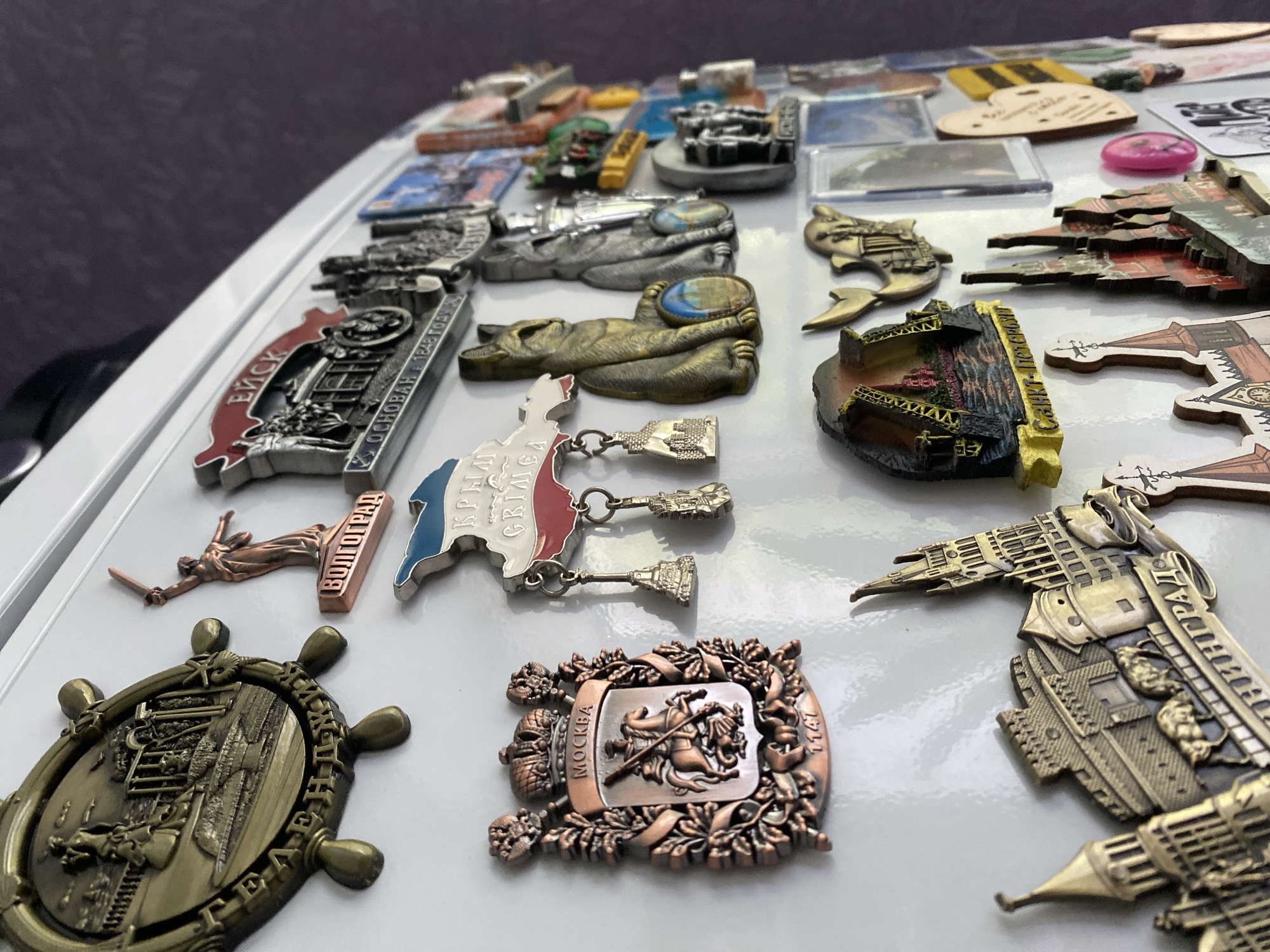 The image displays a collection of refrigerator magnets attached to a white surface, likely a refrigerator door. The magnets are predominantly metallic, with some featuring colored enamel. They depict various landmarks and symbols, predominantly Russian.
On the left, a large, circular magnet with a ship's wheel design is visible, with Cyrillic text around the rim. To its right, a smaller magnet depicts a statue of a woman holding a sword, with the word "ВОЛГОГРАД" (Volgograd) below it. Above this, another magnet shows a historical-looking building and the text "ЕЙСК" (Yeysk) and "ОСНОВАН В 1848 ГОДУ" (Founded in 1848). A magnet shaped like the Crimean peninsula with blue and red borders is also present.
Further to the right, several magnets feature architectural representations. One notable magnet is a crest with a horseman, bearing the inscription "МОСКВА" (Moscow) and the year "1147". Adjacent to this are two magnets shaped like seated cats holding small oval-shaped images. Below these, a magnet depicts a cityscape with intricate buildings and the text "САНКТ-ПЕТЕРБУРГ" (Saint Petersburg). Other magnets show various architectural structures, including towers and cathedrals, some in metallic tones and others colored. There is also a heart-shaped magnet with yellow and black stripes, and a pink circular object which appears to be another magnet.
The background is a blurred purple texture, suggesting a wall or a similar surface behind the refrigerator. There are no people or animals depicted in the scene, beyond the representations on the magnets. The lighting is bright, casting subtle shadows on the white surface, indicating a well-lit indoor environment. The specific location of Lomonosov, Russia, is not explicitly indicated by any of the visible text or imagery on the magnets.