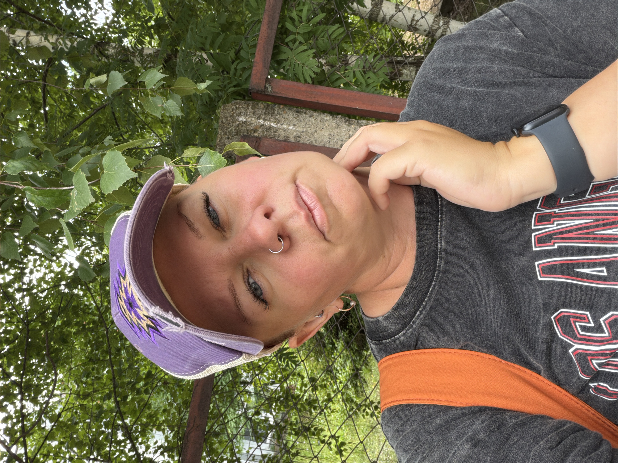 A woman with short blonde hair and a nose ring is taking a selfie outdoors, wearing a purple baseball cap and a dark t-shirt with an orange strap from a backpack visible over her shoulder. She is resting her chin on her hand. In the background, there is a fence, trees, and a concrete pillar. The location is the city of Chaikovsky, Russia.
